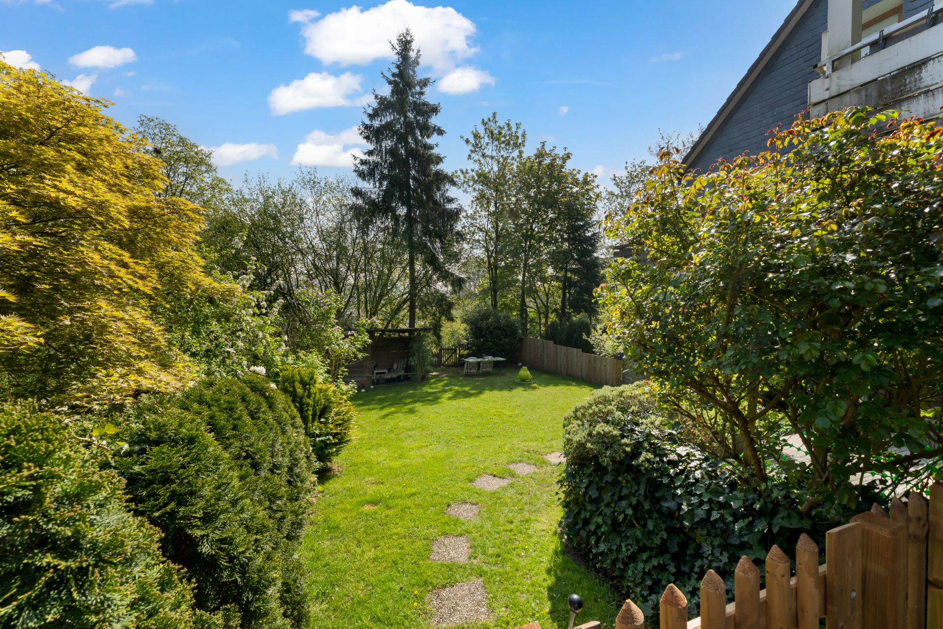 Grüner Garten mit Rasen, Bäumen, Sträuchern und einem kleinen Holzzaun im Vordergrund.