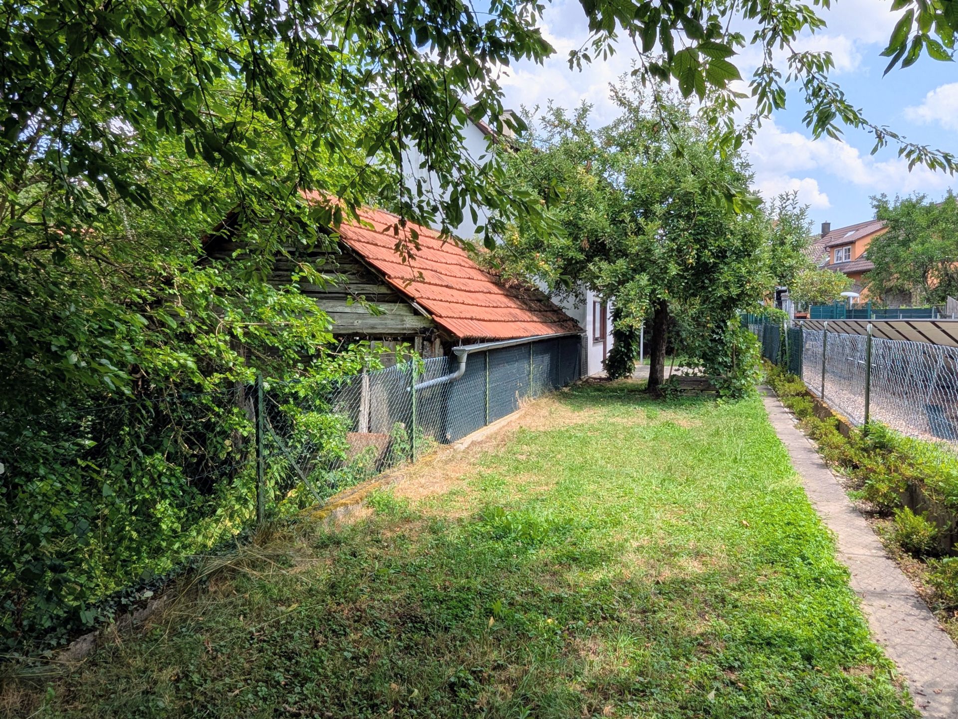 Garten mit Rasen, Zaun, Bäumen und einem Gebäude mit rotem Dach im Hintergrund.