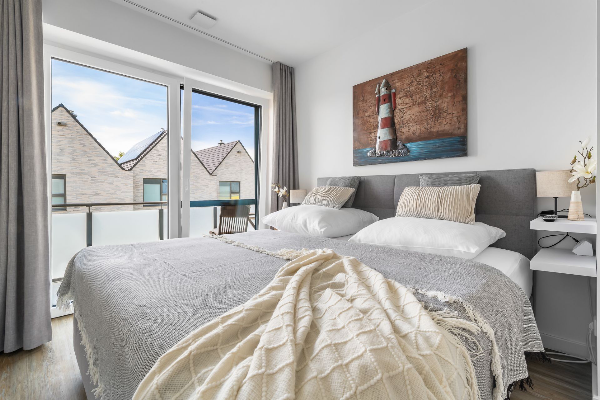 Modernes Schlafzimmer mit Doppelbett, großem Fenster und Bild eines Leuchtturms an der Wand.