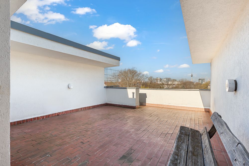 Große Terrasse mit roten Fliesen, weißen Wänden und Holzbänken, Blick auf den Himmel.
