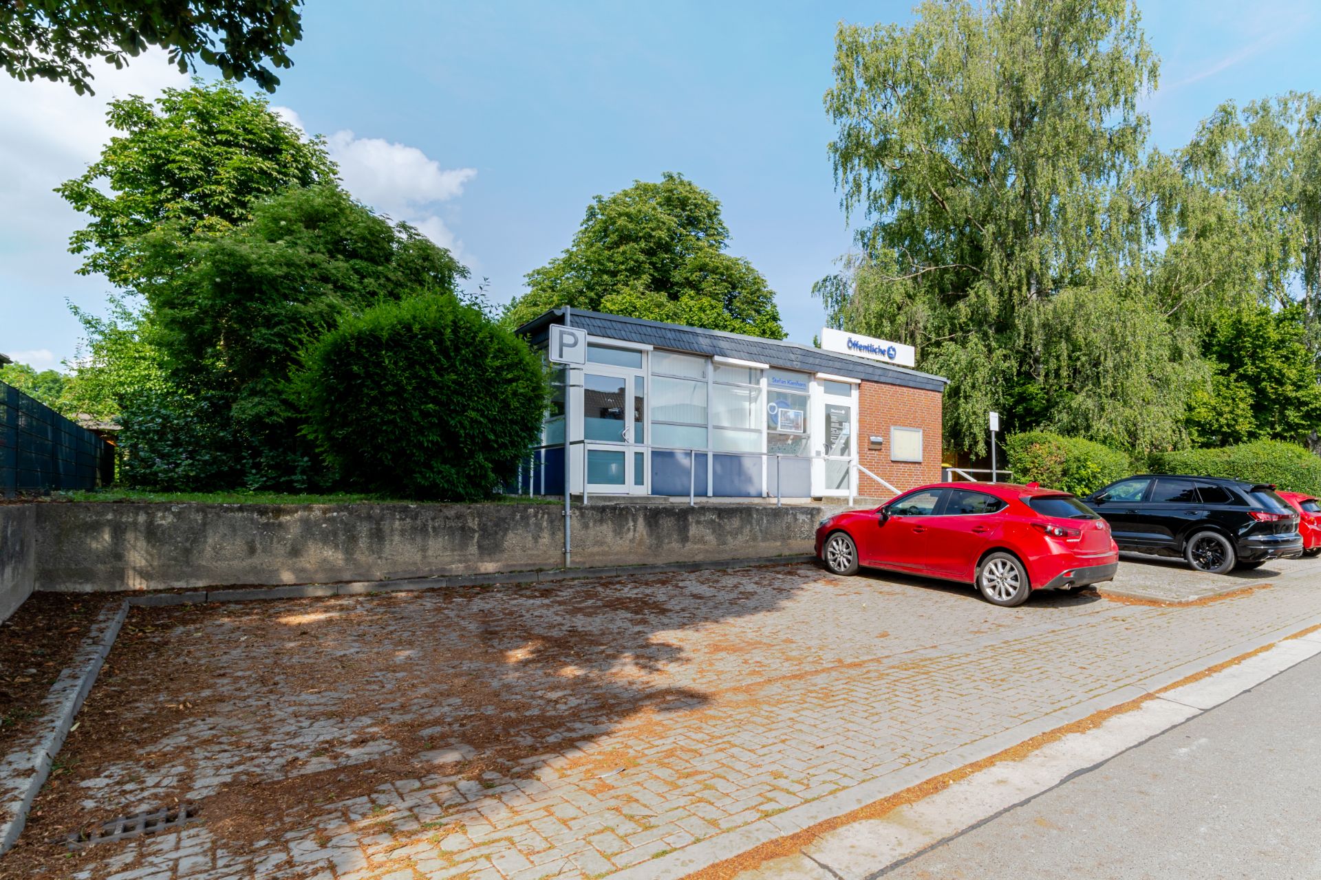 Kleines Gebäude mit großen Fenstern und Parkplatz, mehrere Autos parken davor.