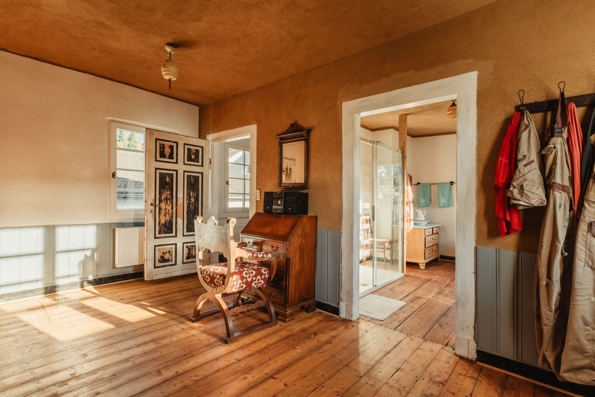 Heller Raum mit Holzboden, Schreibtisch, Sesseln und Garderobe. Im Hintergrund ein Bad mit Dusche.