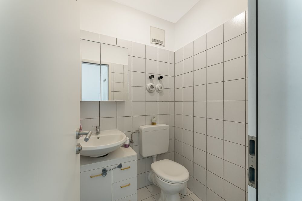 Kleines Badezimmer mit Waschbecken, Spiegel und Toilette, weiße Fliesen an den Wänden.