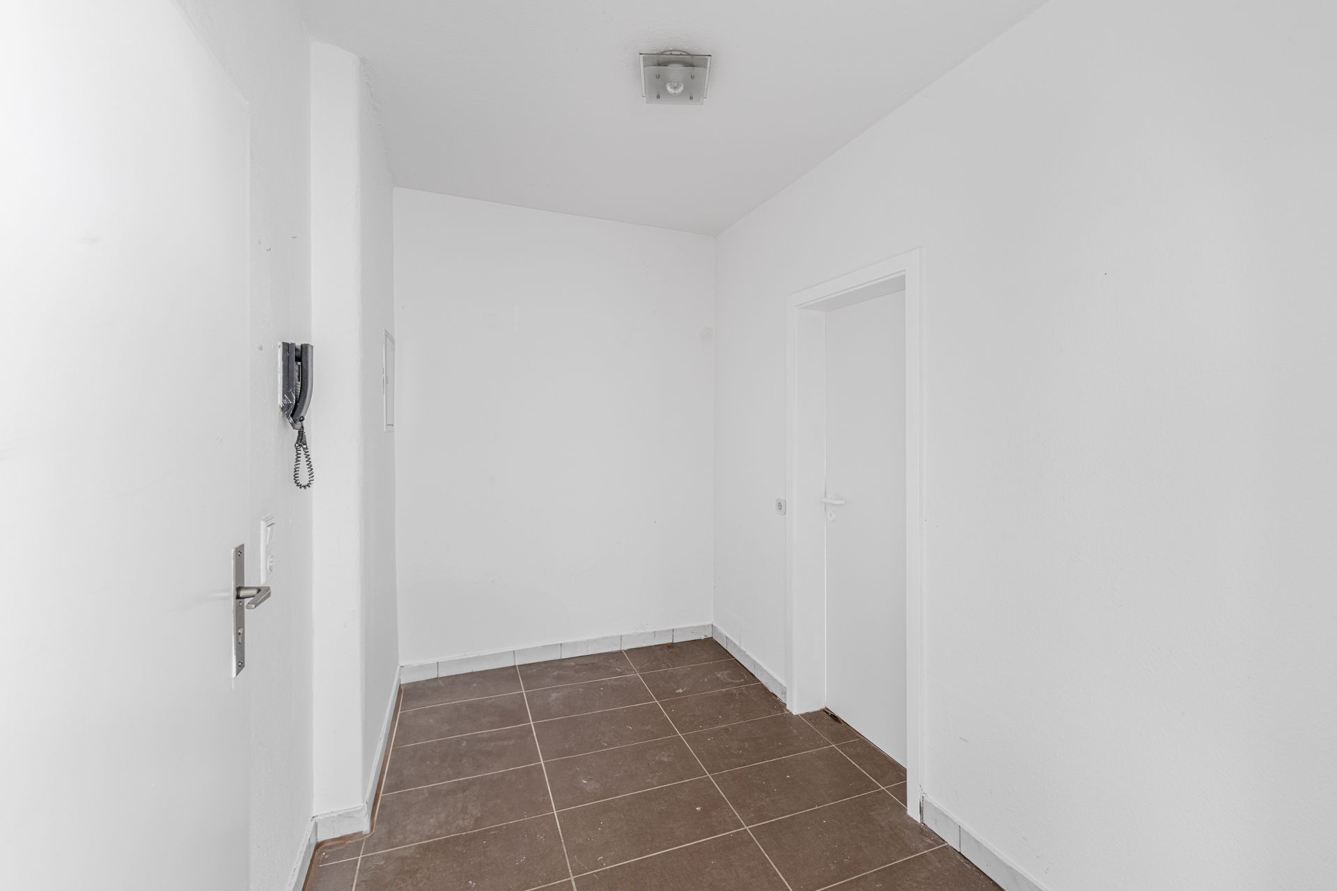Weißer Flur mit braunen Fliesen, zwei Türen und einer Gegensprechanlage an der Wand.