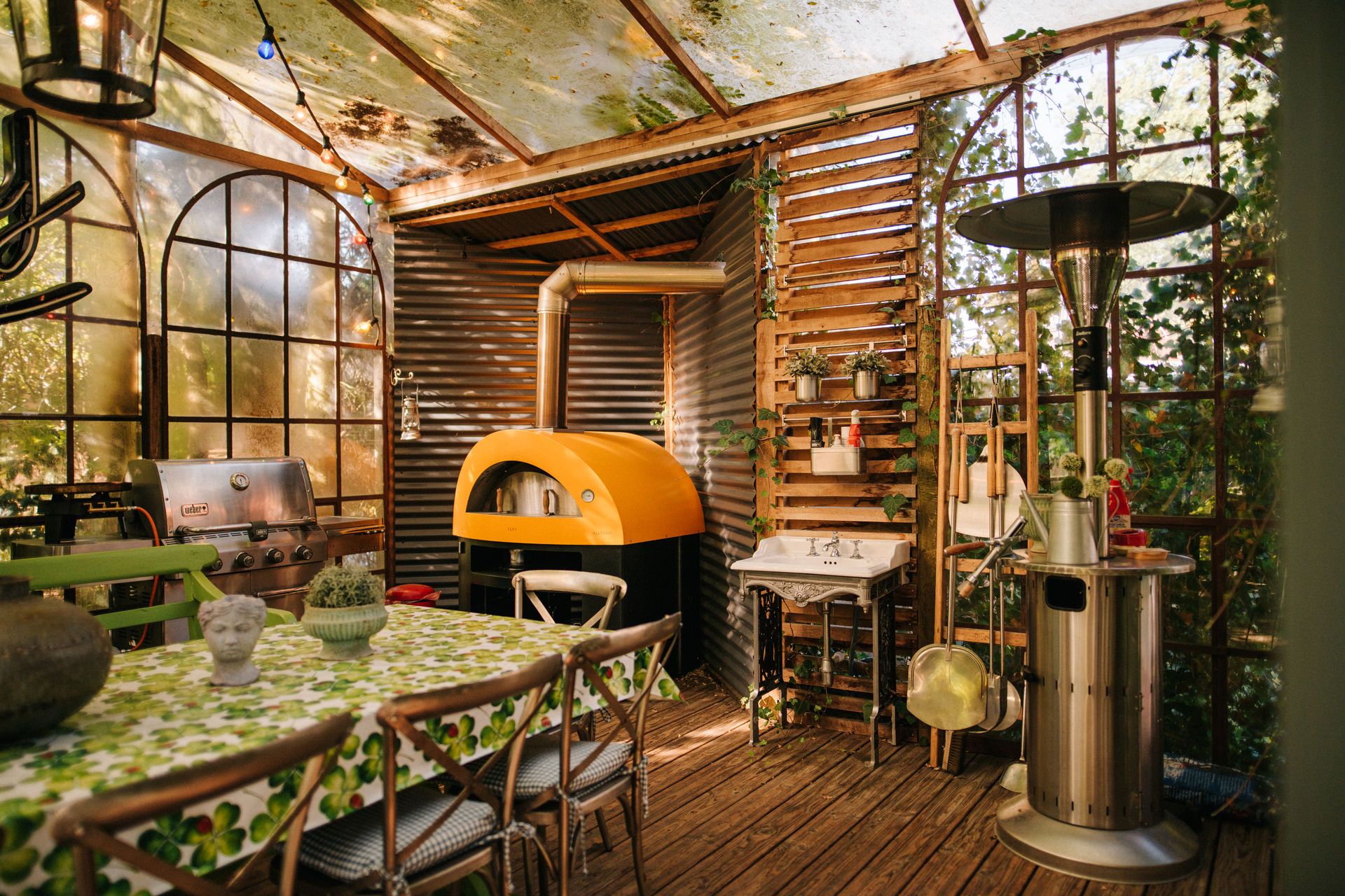 Überdachte Terrasse mit Esstisch, Holzofen, Grill und Heizstrahler, viele Pflanzen sichtbar.