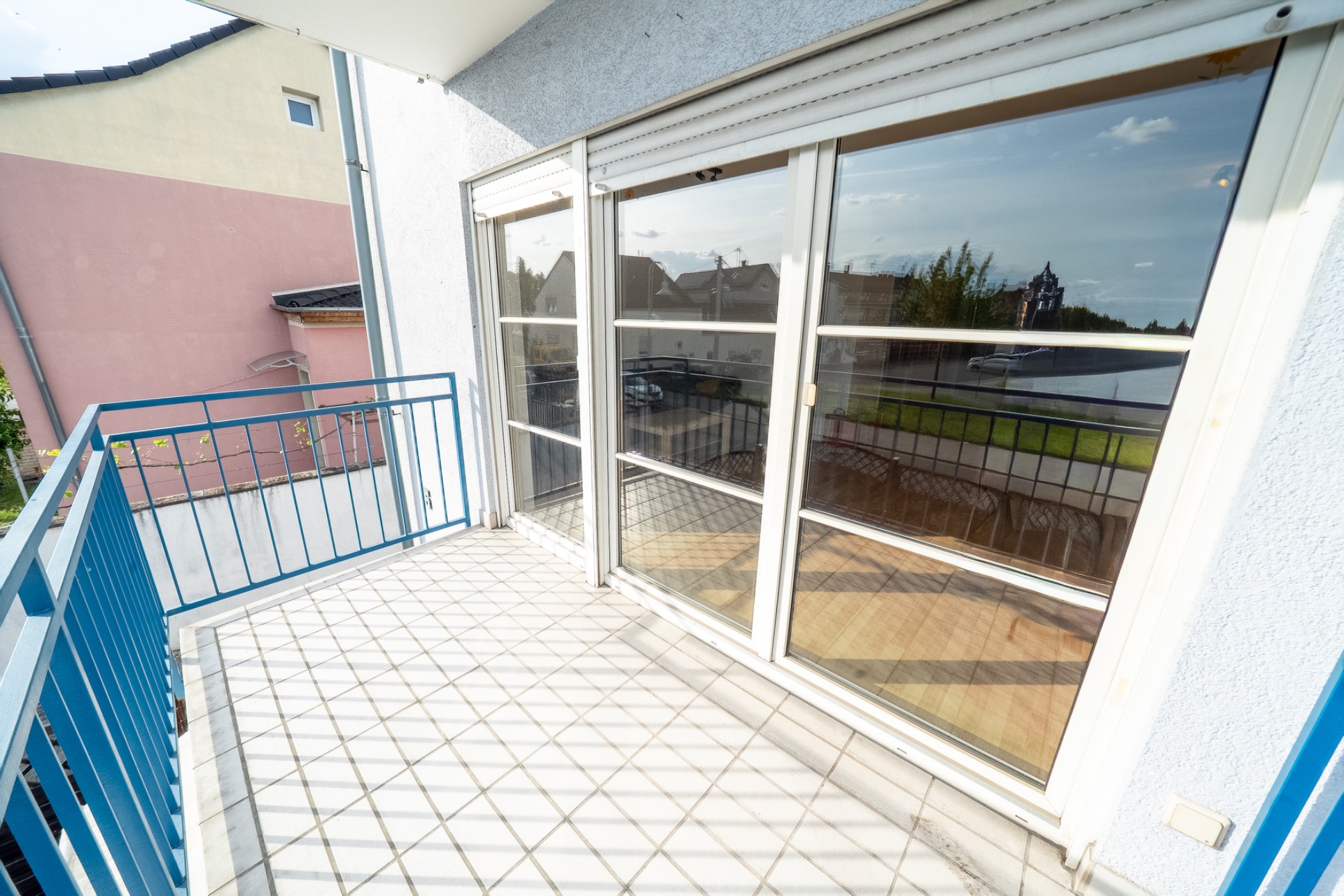 Heller Balkon mit blauen Geländern und großen Fenstern, Blick auf Nachbargebäude und Straße.