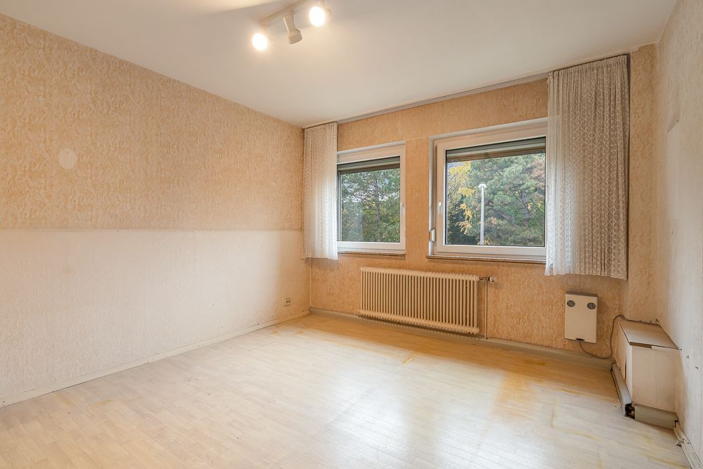 Leeres Zimmer mit zwei Fenstern, Vorhängen und Heizkörper, helle Wände und Holzboden.