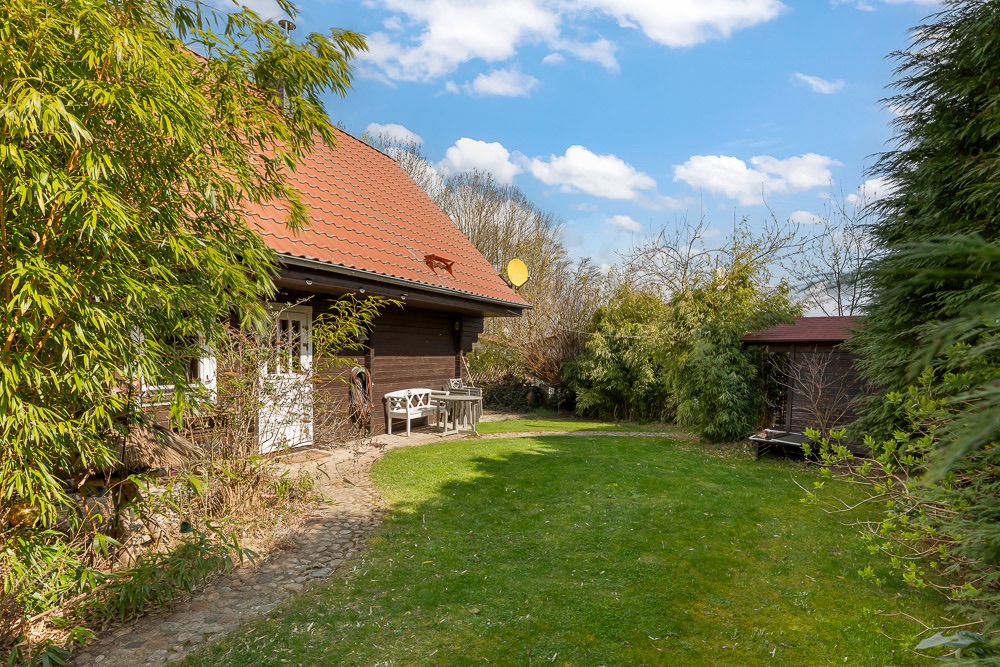 Haus mit rotem Dach und Garten, umgeben von Bäumen und Büschen, eine Bank vor dem Eingang.