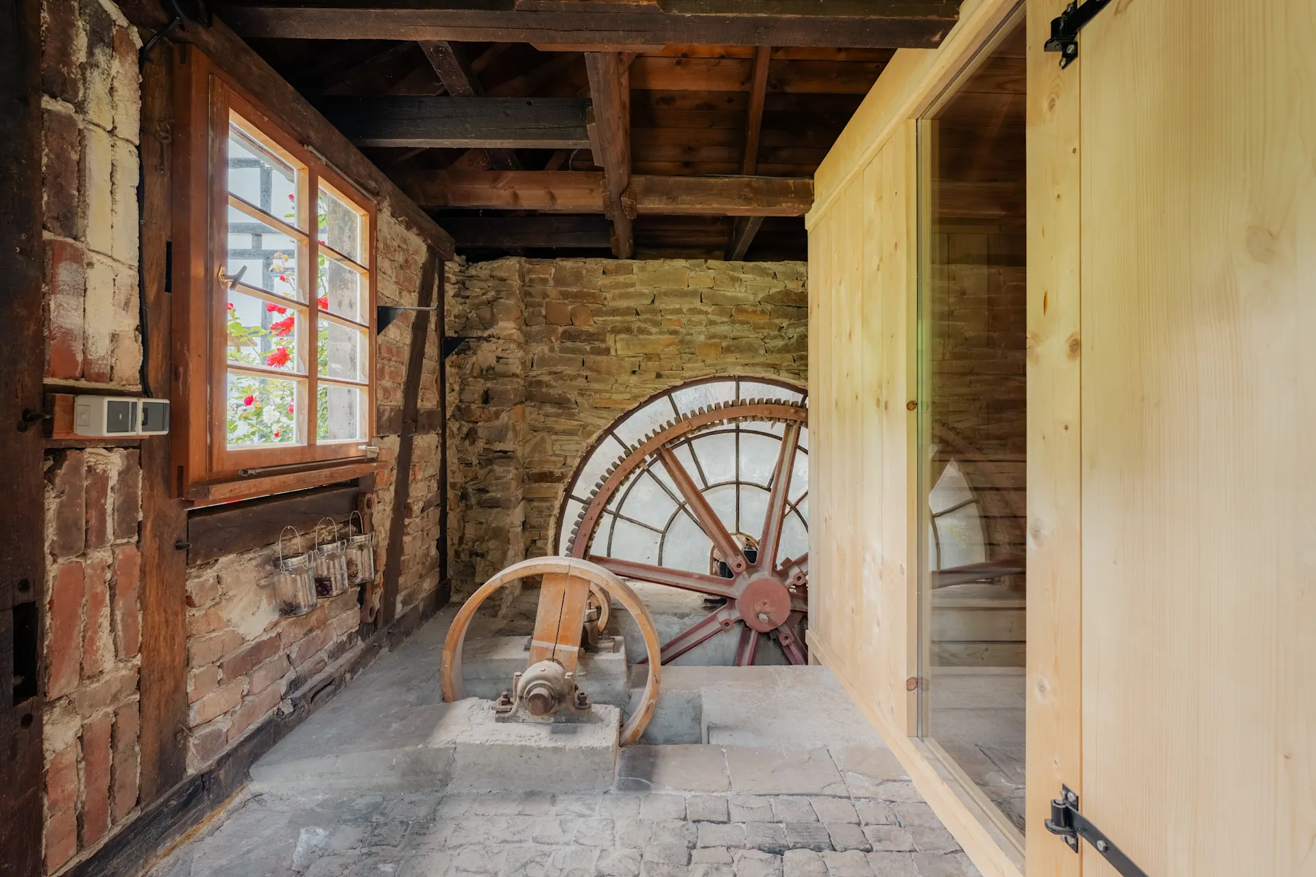 Raum mit Holzbalkendecke, Steinwand und großem historischen Mühlrad aus Metall.
