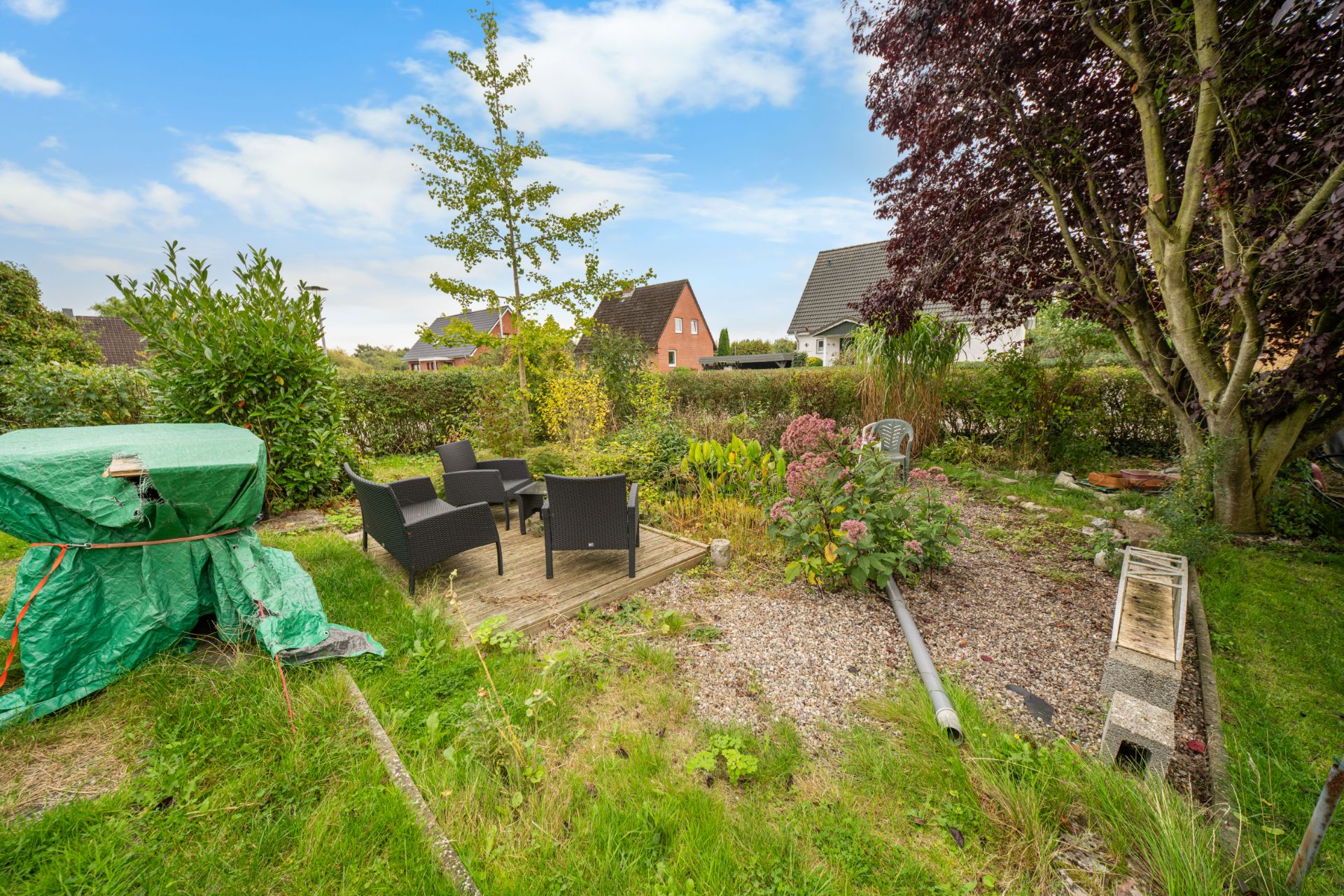 Garten mit Holzdielen, Gartenmöbeln und vielen Pflanzen, daneben ein mit Plane abgedeckter Gegenstand.