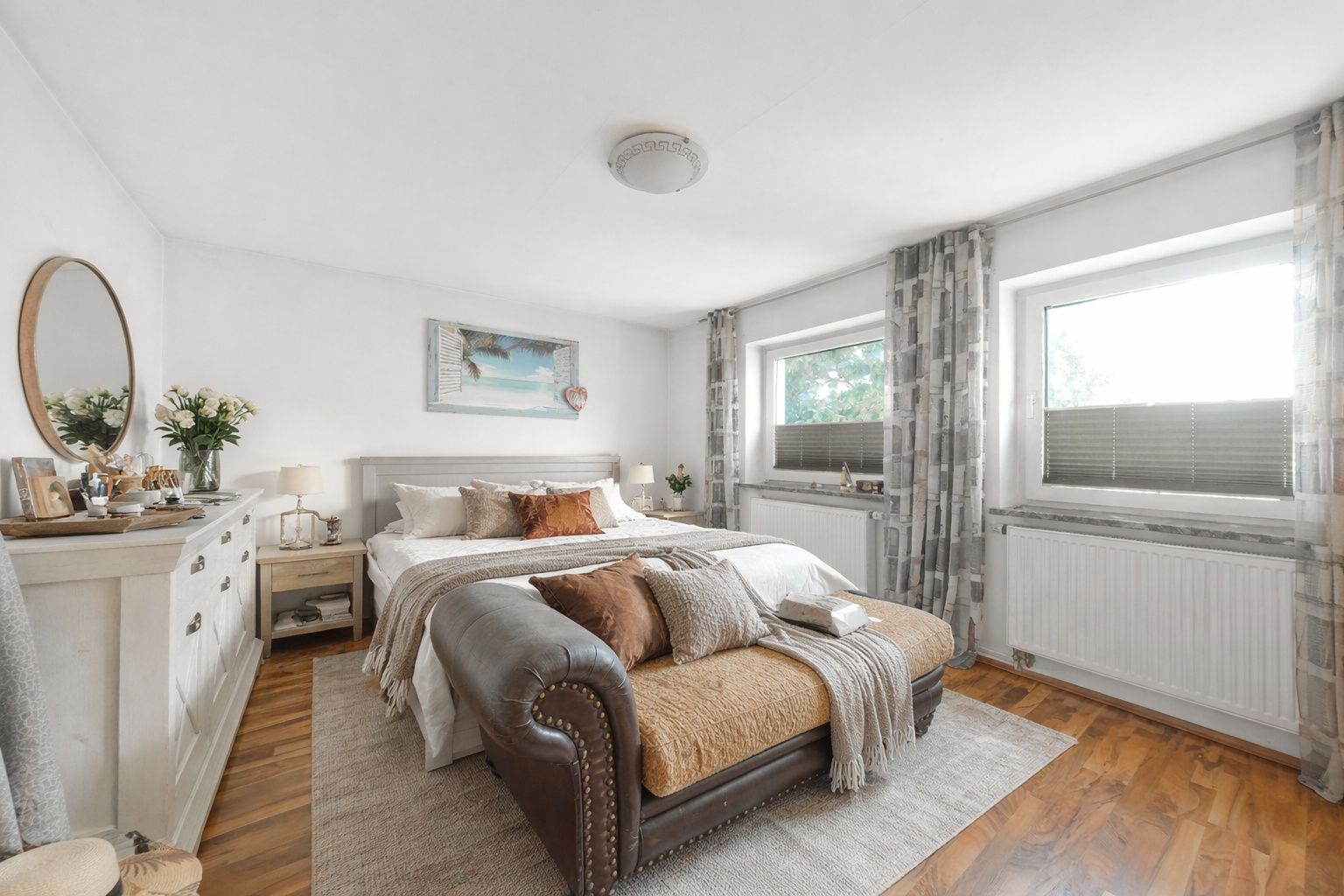 Helles Schlafzimmer mit großem Bett, Sofa am Fußende, zwei Fenstern und Holzboden.