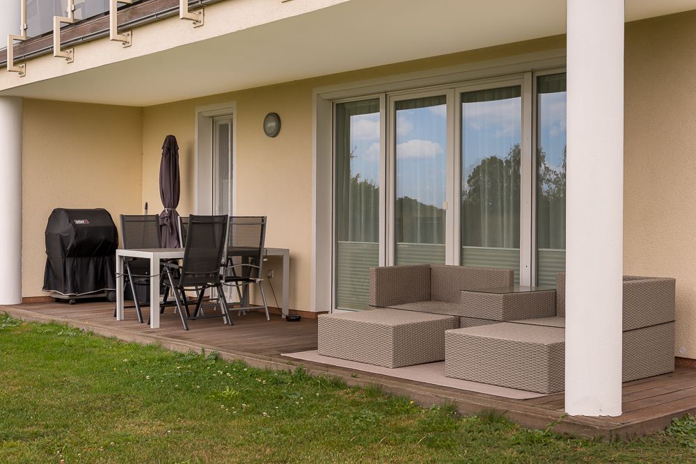 Überdachte Terrasse mit Sitzgarnitur, Esstisch, Stühlen und Grill, angrenzend an Rasenfläche.