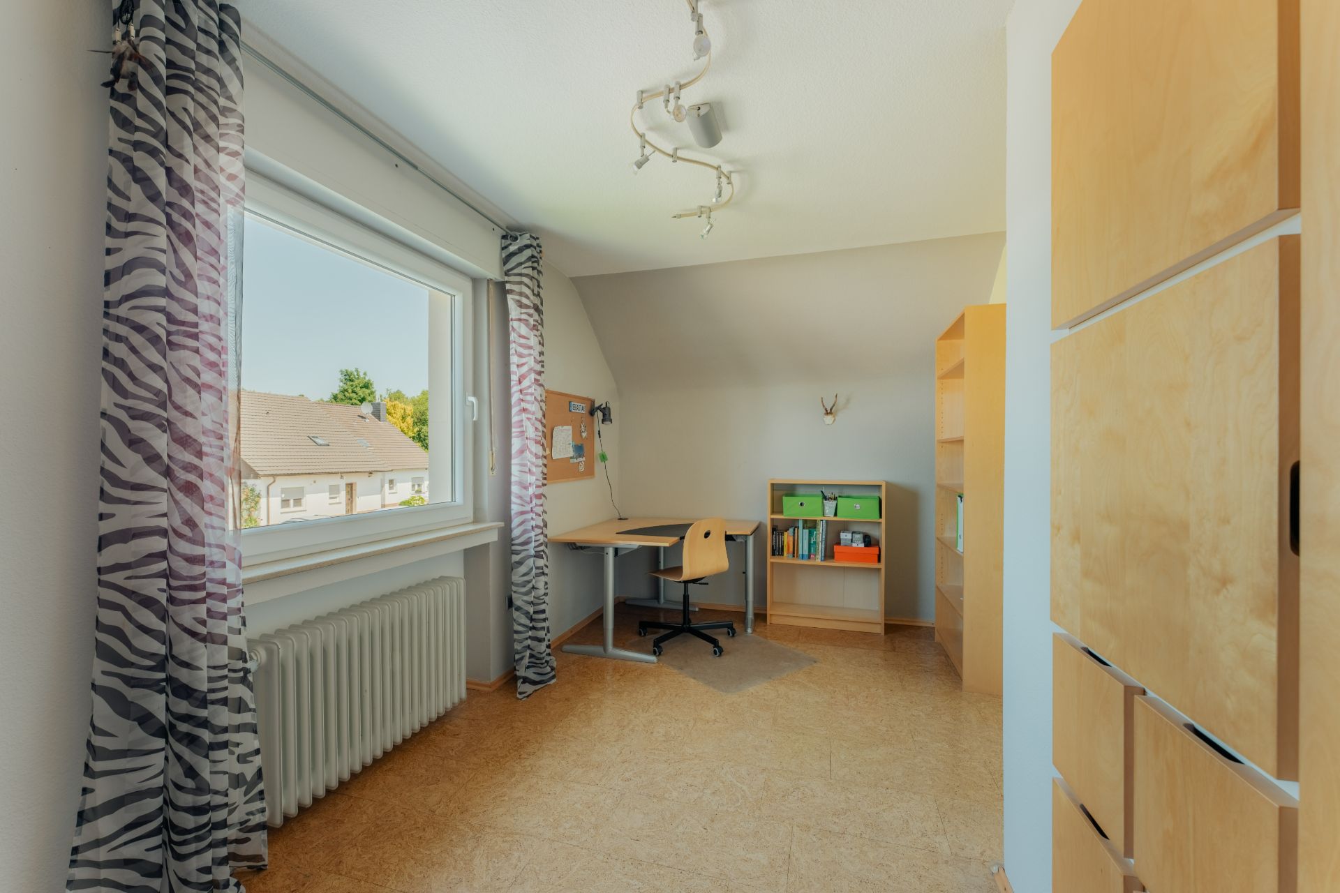 Helles Arbeitszimmer mit großem Fenster, Schreibtisch, Regal und Stuhl.