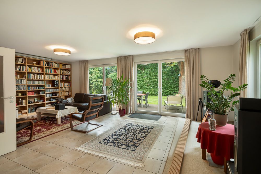 Helles Wohnzimmer mit Bücherregal, Teppich, Sesseln und Blick auf den Garten durch große Glasfenster.