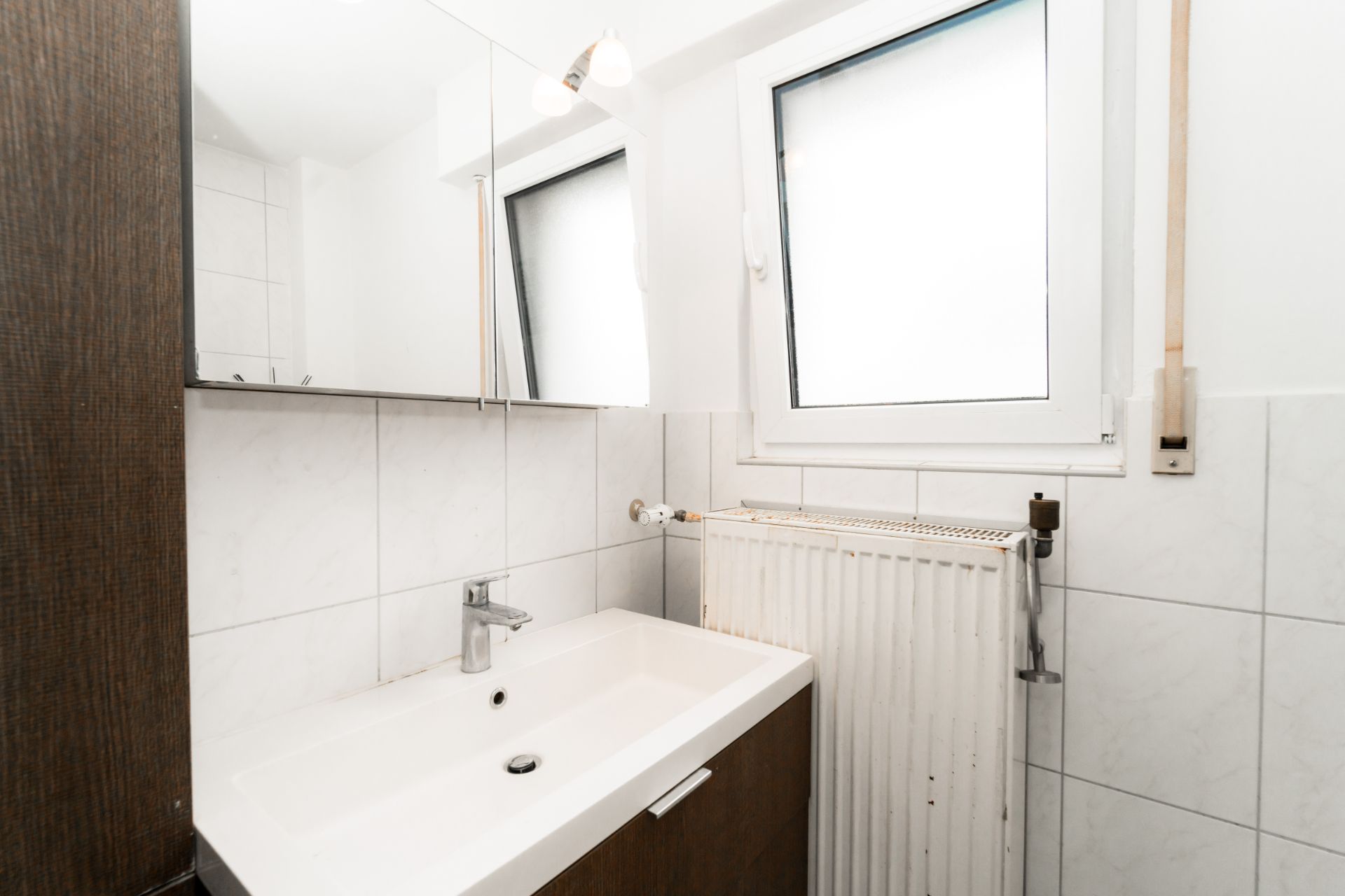 Kleines Badezimmer mit Waschbecken, Spiegel, Heizkörper und Fenster.