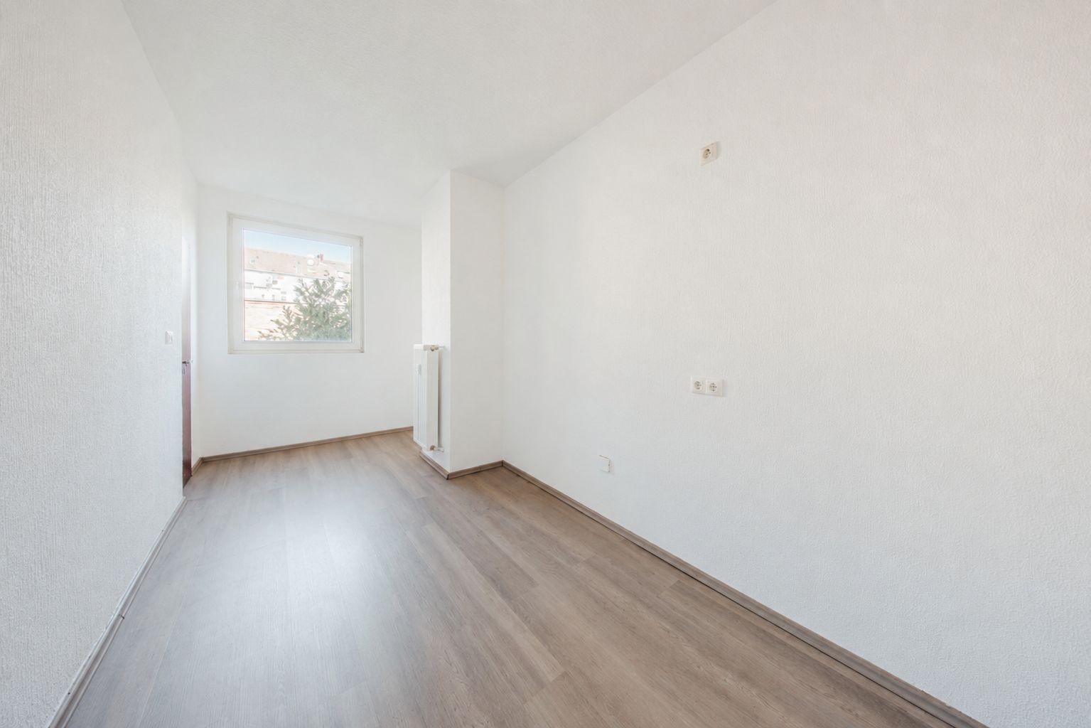 Leerer, heller Raum mit weiß gestrichenen Wänden, großem Fenster und hellem Holzboden.