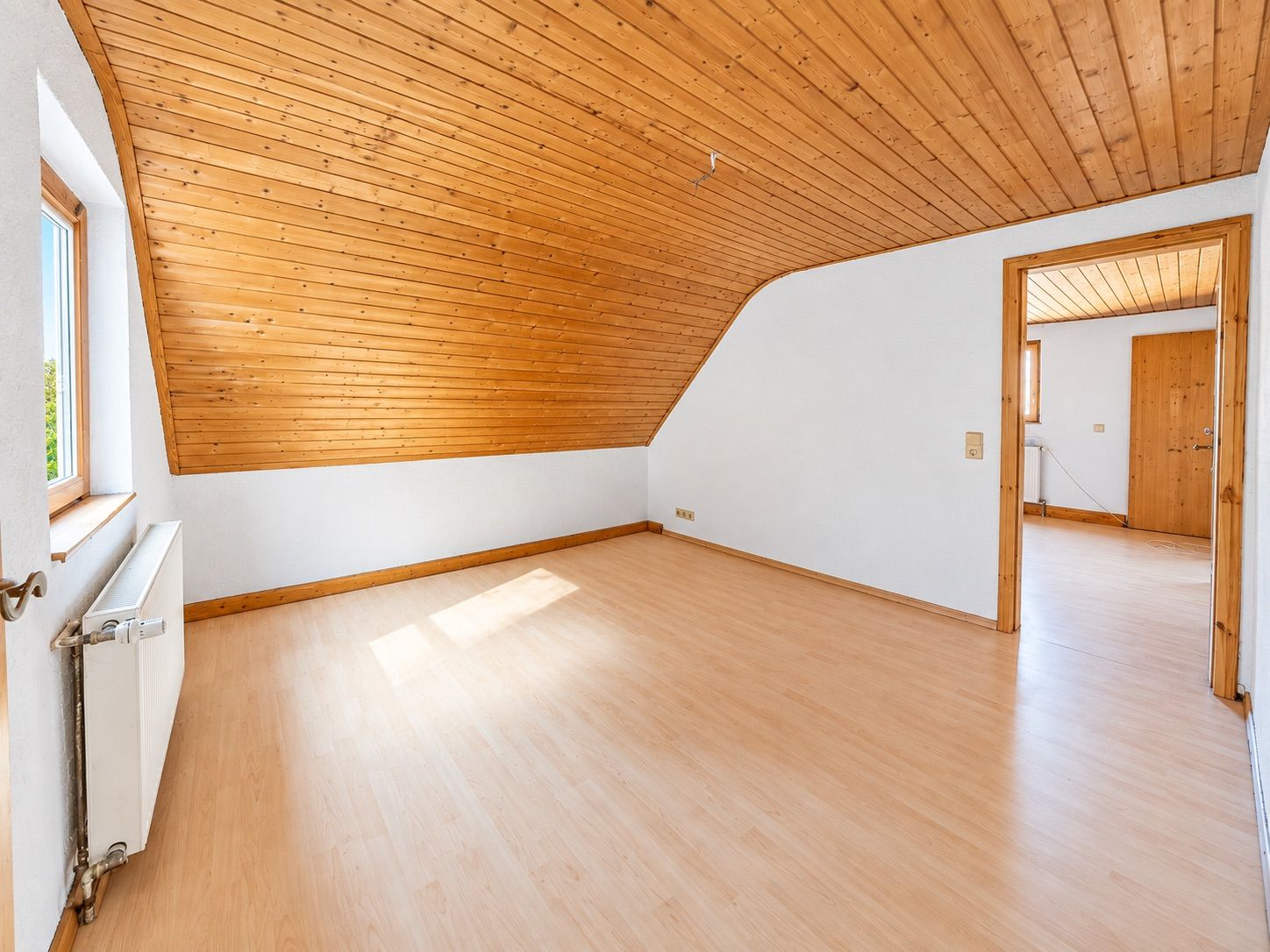 Helles Zimmer mit Holzdecke, Holzfußboden, Fenster und weiß gestrichenen Wänden.