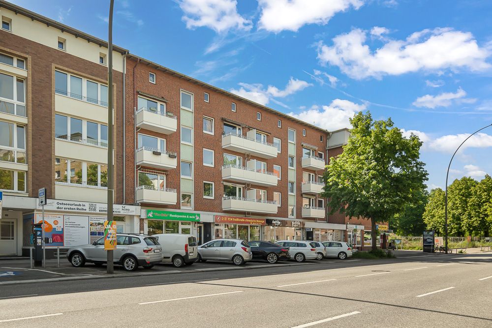 Mehrstöckiges Wohnhaus mit Balkonen und geparkten Autos an der Straße, Bäume und blauer Himmel im Hintergrund.