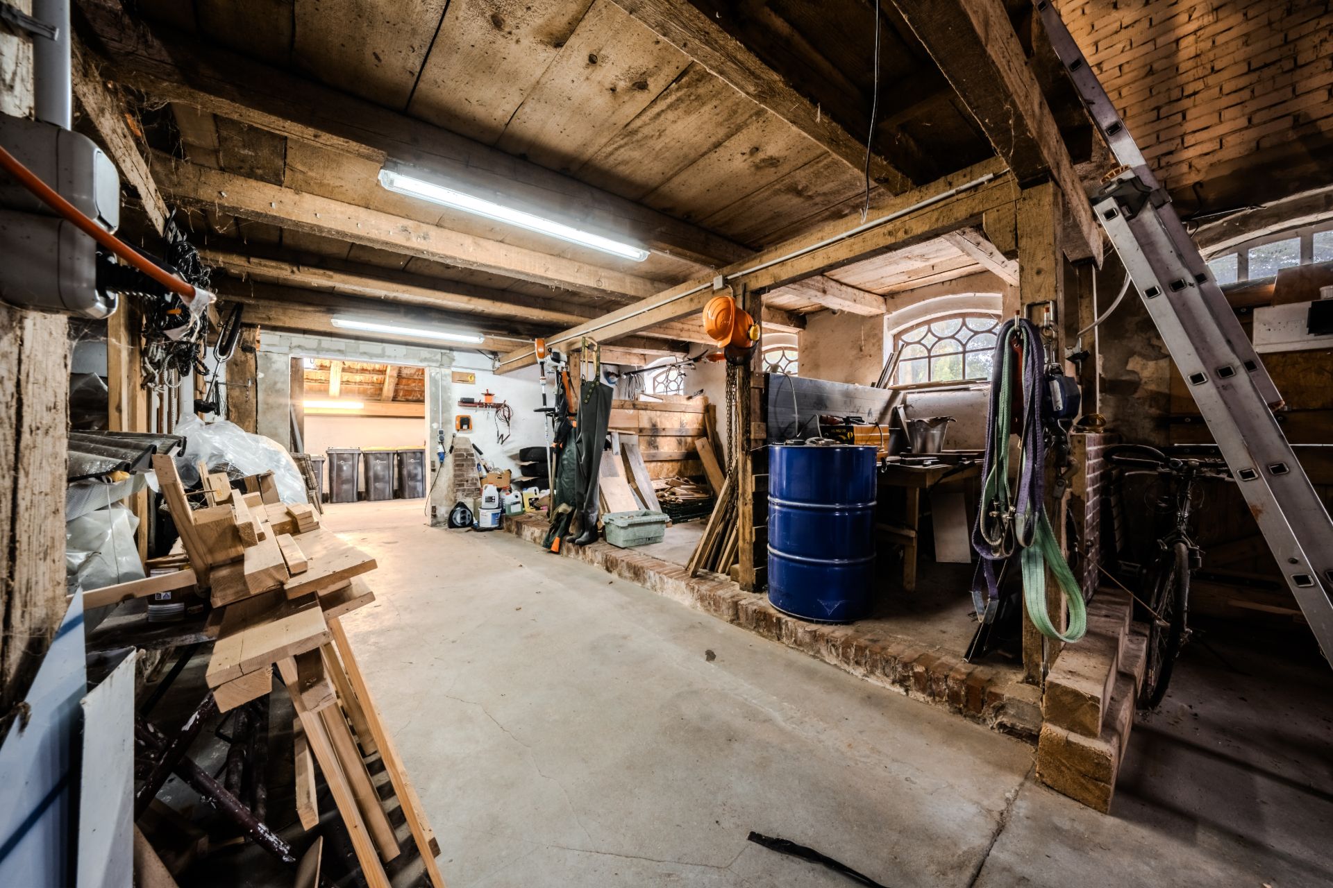 Geräumige Werkstatt mit Holzregalen, Werkzeugen, blauer Tonne und Leiter, rustikale Wände und Decke.