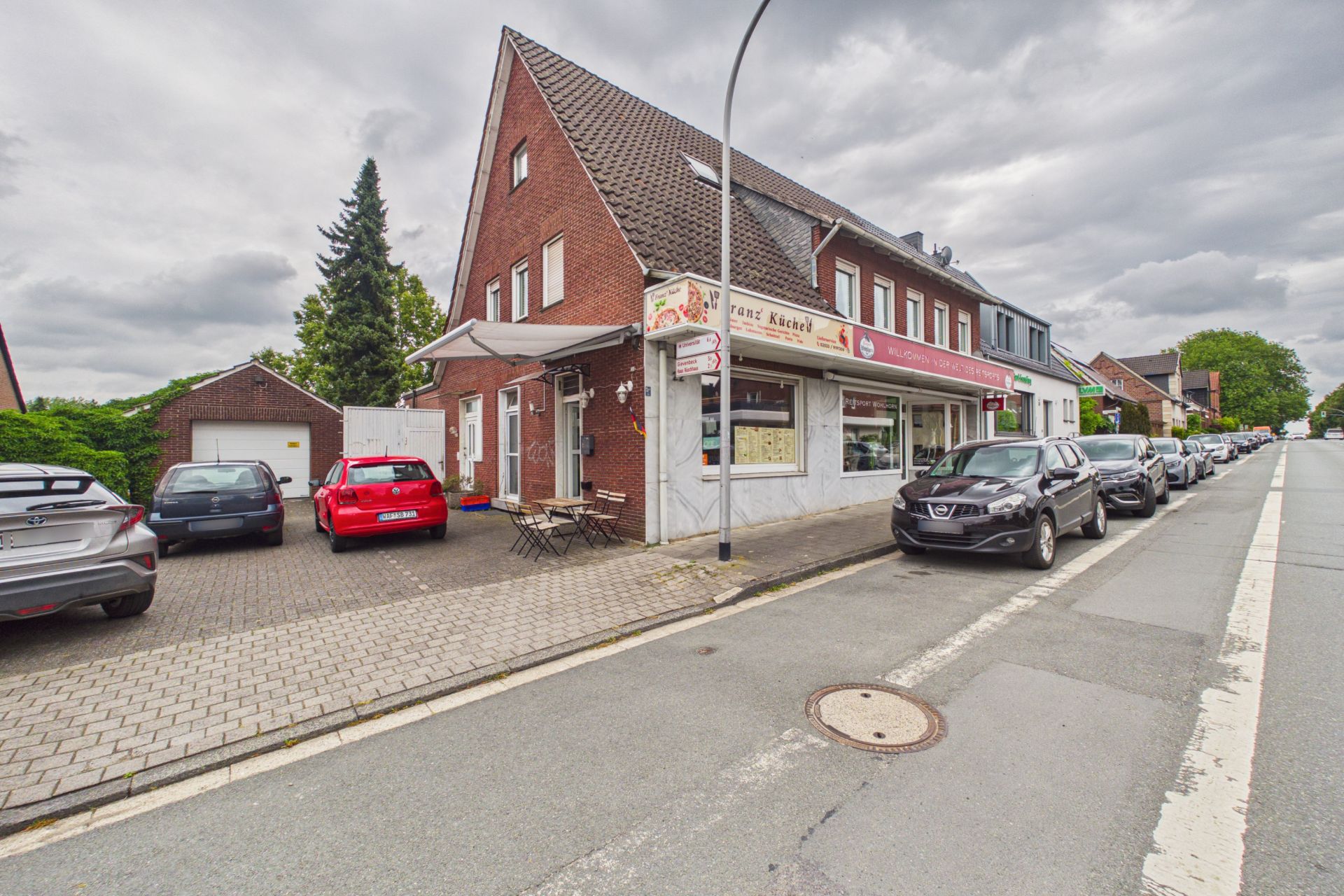 Backsteinwohnhaus mit Laden im Erdgeschoss, parkende Autos und Straße im Vordergrund.