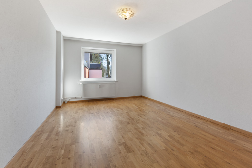 Leerer Raum mit hellem Holzboden, weißer Wand und Fenster mit Heizkörper darunter.