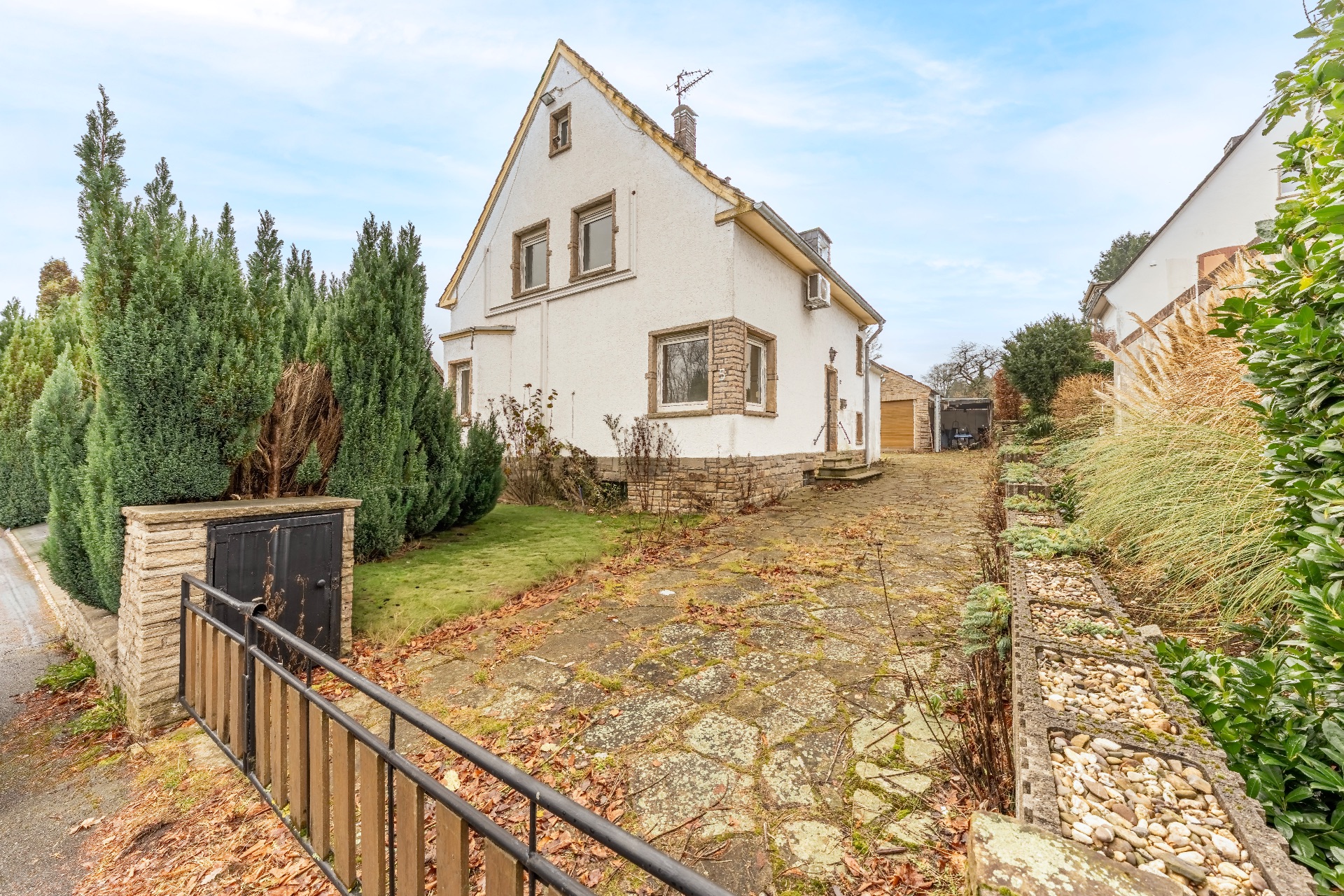 Weißes Einfamilienhaus mit steilem Dach, Einfahrt und Garten, von Hecken und Pflanzen umgeben.