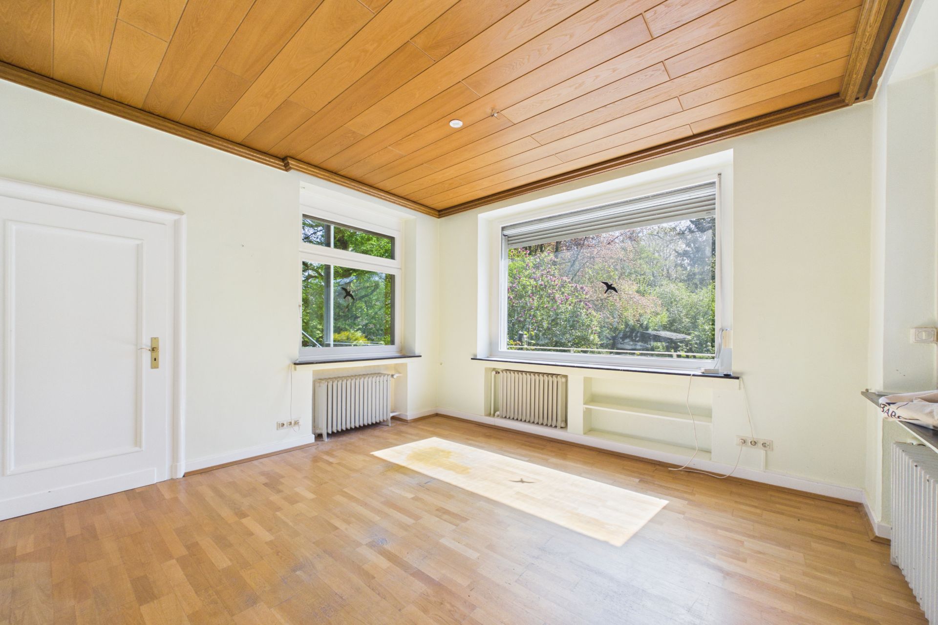 Helles, leeres Zimmer mit Holzdecke, Parkettboden und großen Fenstern mit Blick ins Grüne.