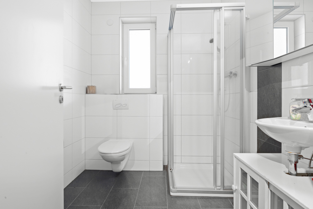 Modernes Badezimmer mit Dusche, WC, Fenster und Waschbecken mit Spiegel.