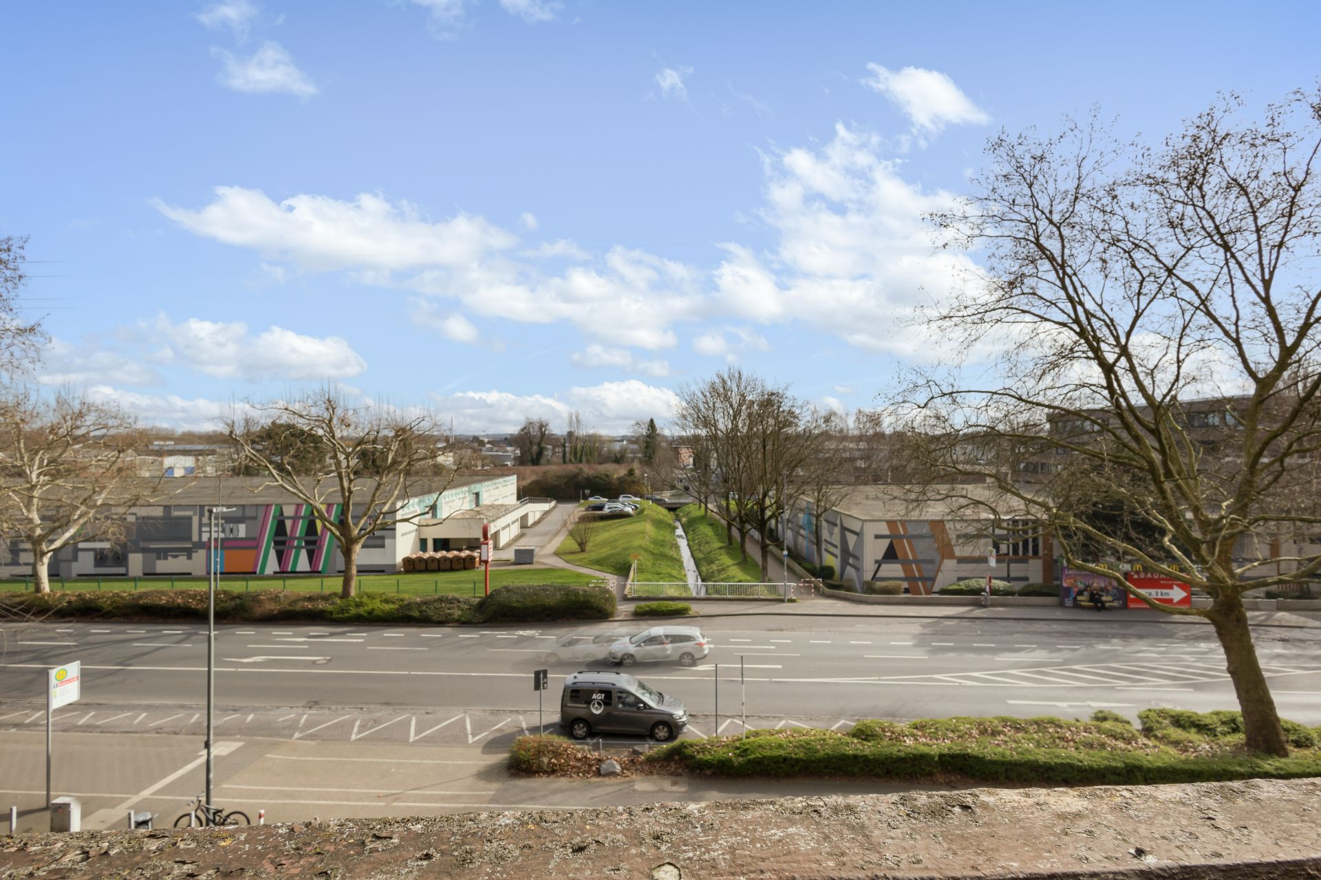 Blick auf eine Straße mit parkenden Autos, Gehweg, Bäumen und Gebäuden im Hintergrund.