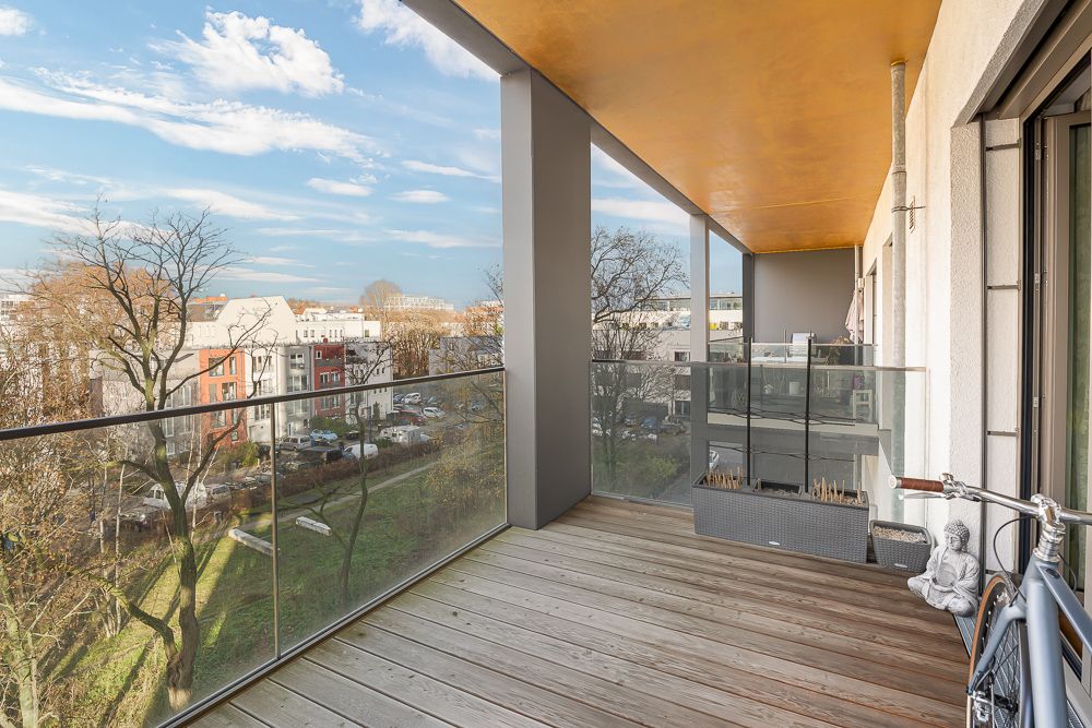 Moderner Balkon mit Holzboden und Glasgeländer, Blick auf Bäume und Wohnhäuser.