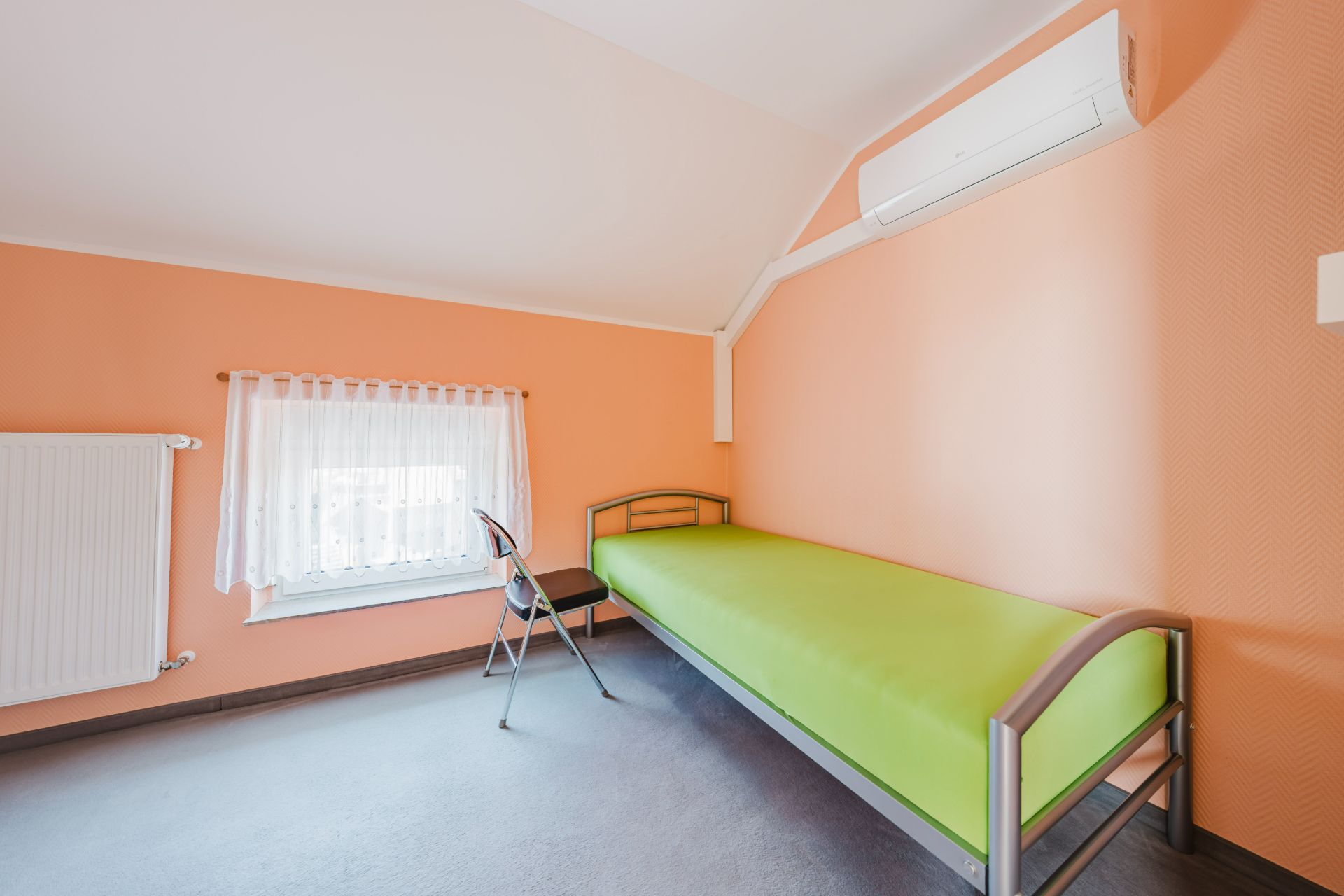 Ein einfaches Zimmer mit grünem Einzelbett, Stuhl, Fenster mit Gardine und Klimaanlage an der Wand.