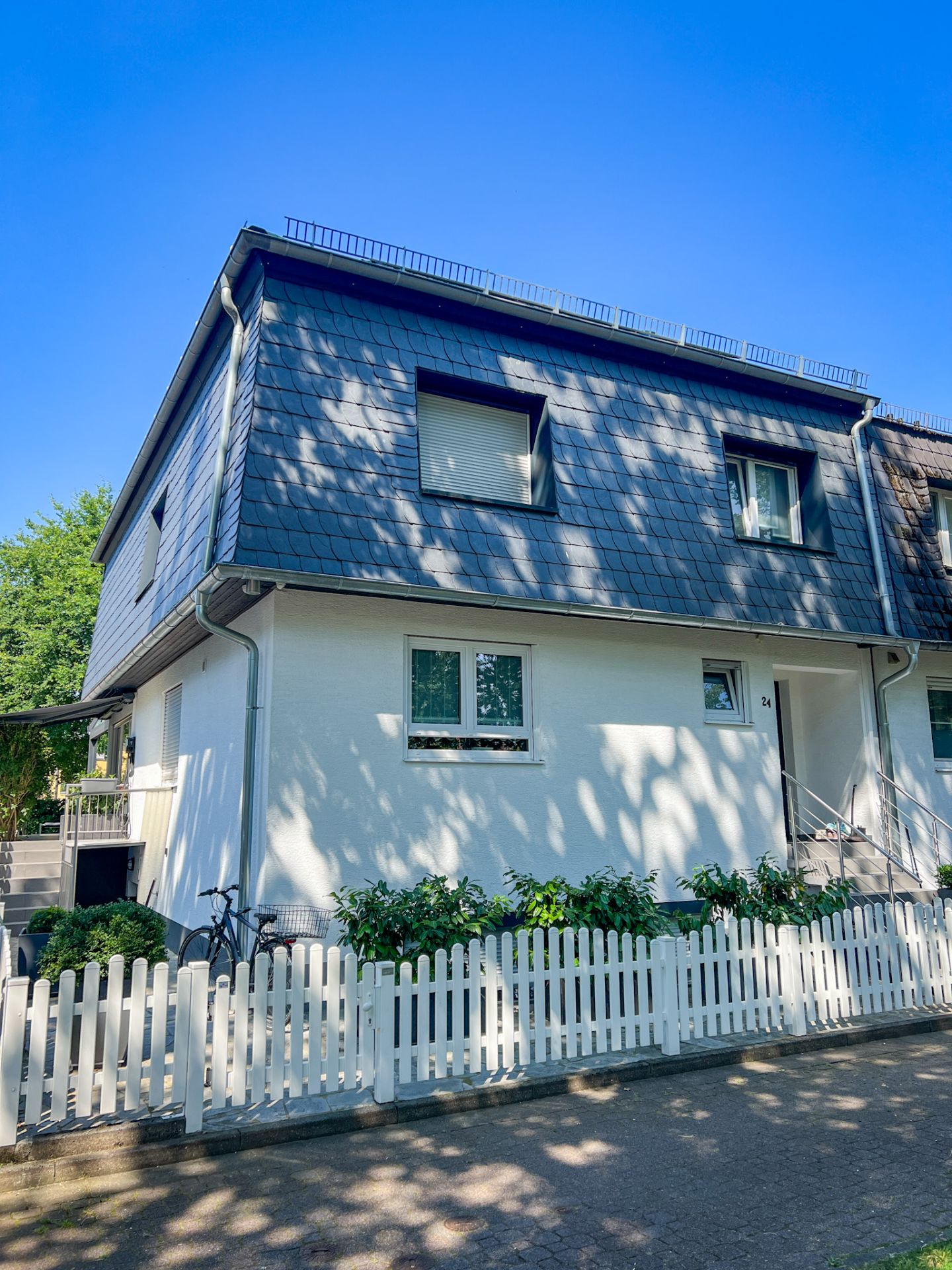 Zweistöckiges Wohnhaus mit dunklem Dach, weißem Zaun und grünen Sträuchern davor.