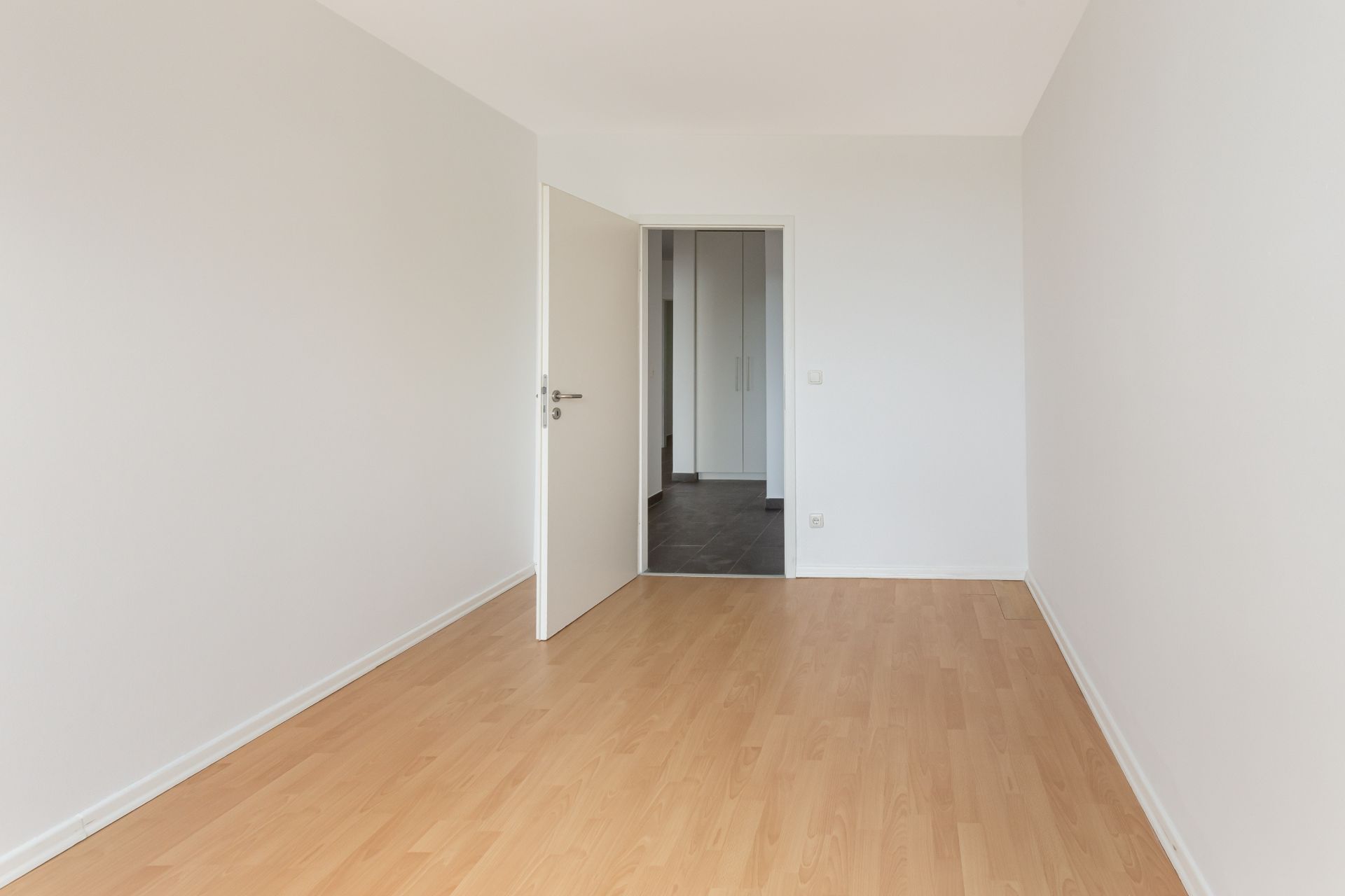Leeres Zimmer mit hellem Holzfußboden, weißen Wänden und geöffneter Tür.