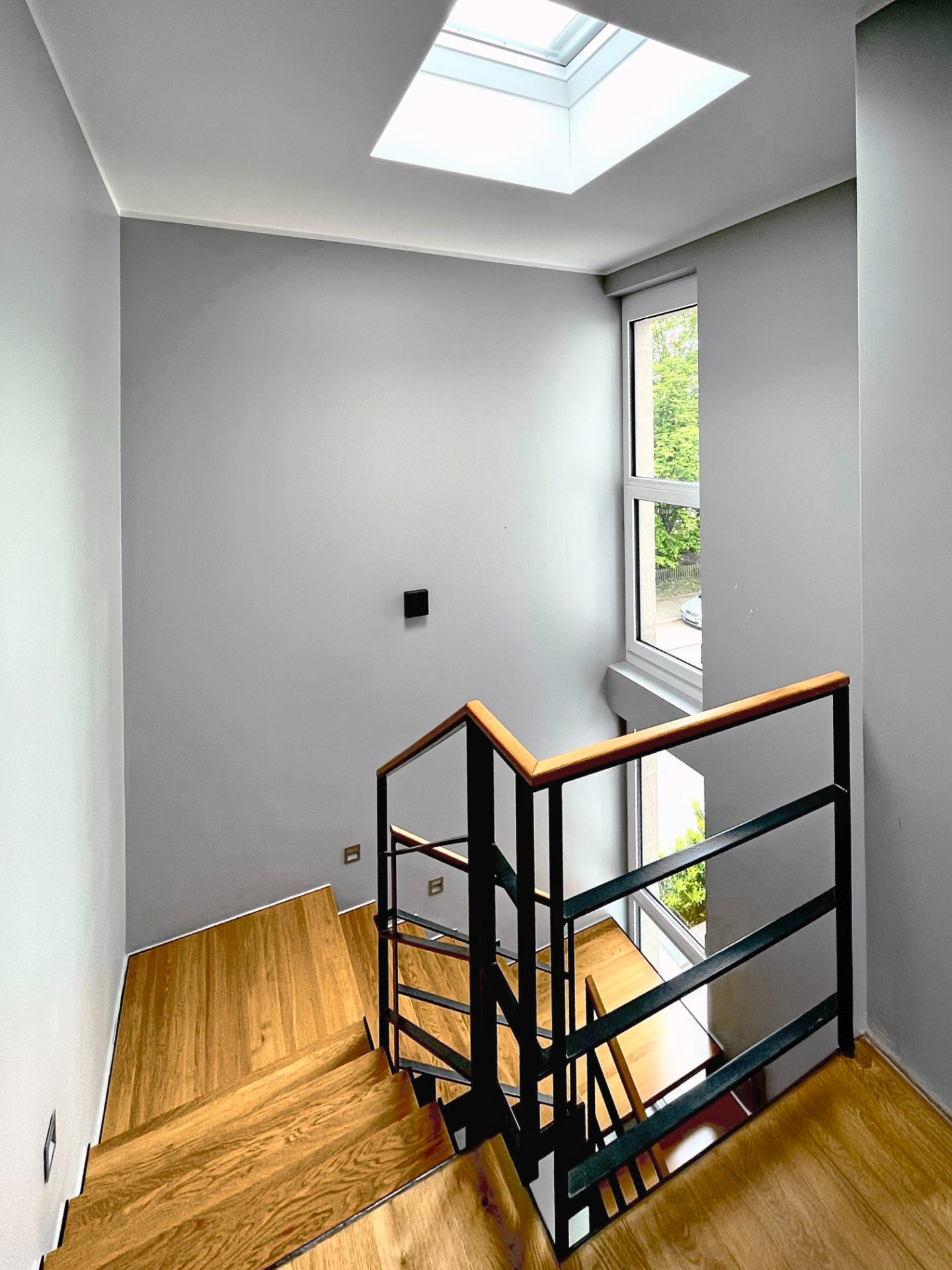 Holztreppe mit schwarzem Geländer, Oberlicht und großem Fenster auf grauer Wand.
