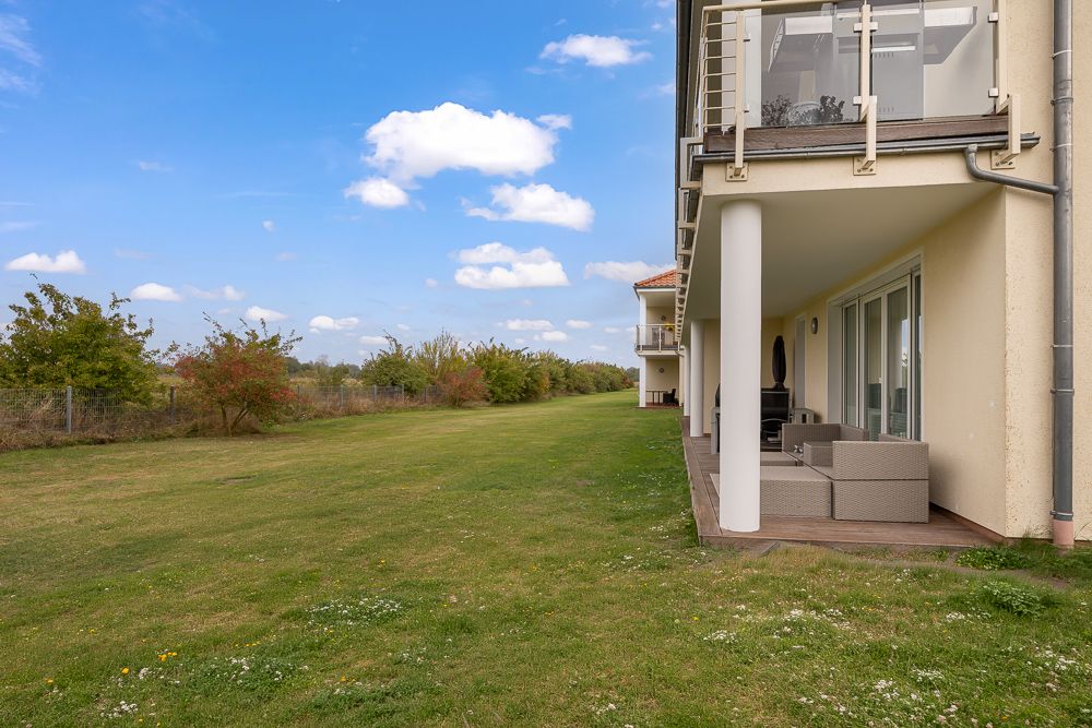Terrasse mit Sitzmöbeln an einem Wohnhaus, großer grüner Rasen und Bäume im Hintergrund.