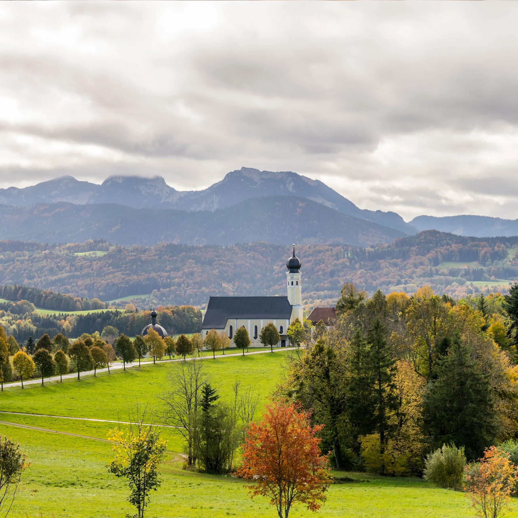 Landschaft mit Kirche, Bäumen und Bergen im Hintergrund unter bewölktem Himmel.