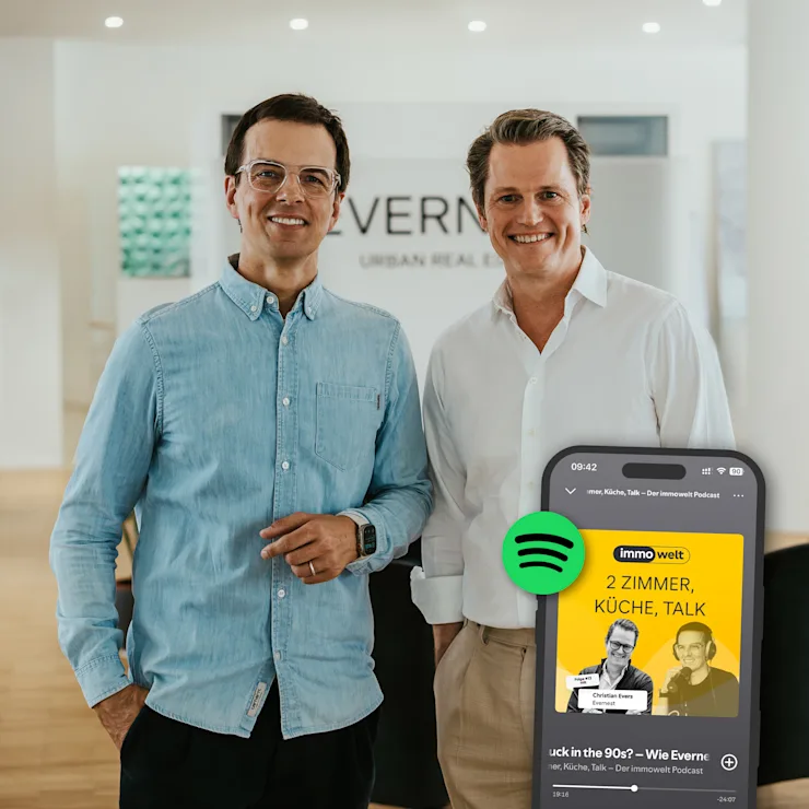 Zwei Männer stehen lächelnd in einem modernen, hellen Raum mit Holzboden.