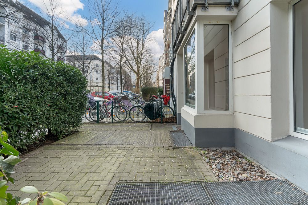 Gepflasterter Weg mit Sträuchern, Fensterfront und mehreren Fahrrädern am Zaun.