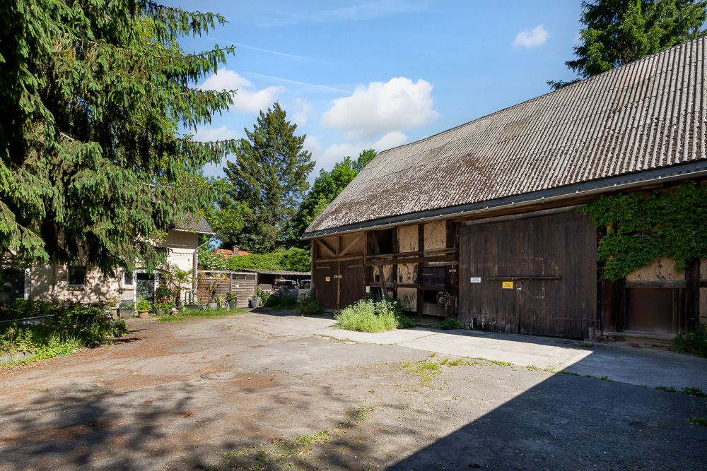Großer Hof mit Scheune aus Holz und Stein, umgeben von Bäumen und Pflanzen.