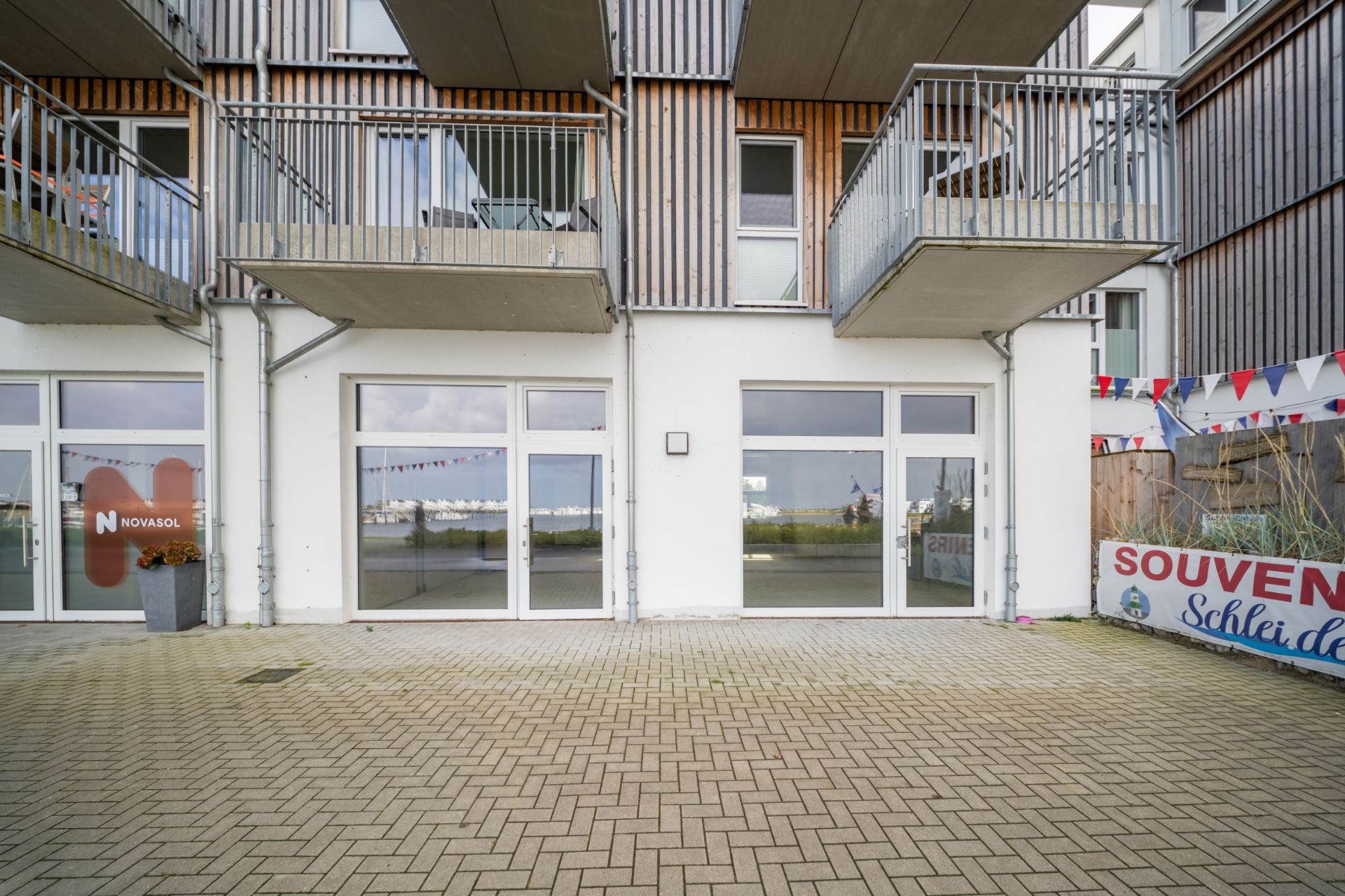 Gebäude mit großen Fenstern, zwei Balkonen und gepflastertem Vorplatz.