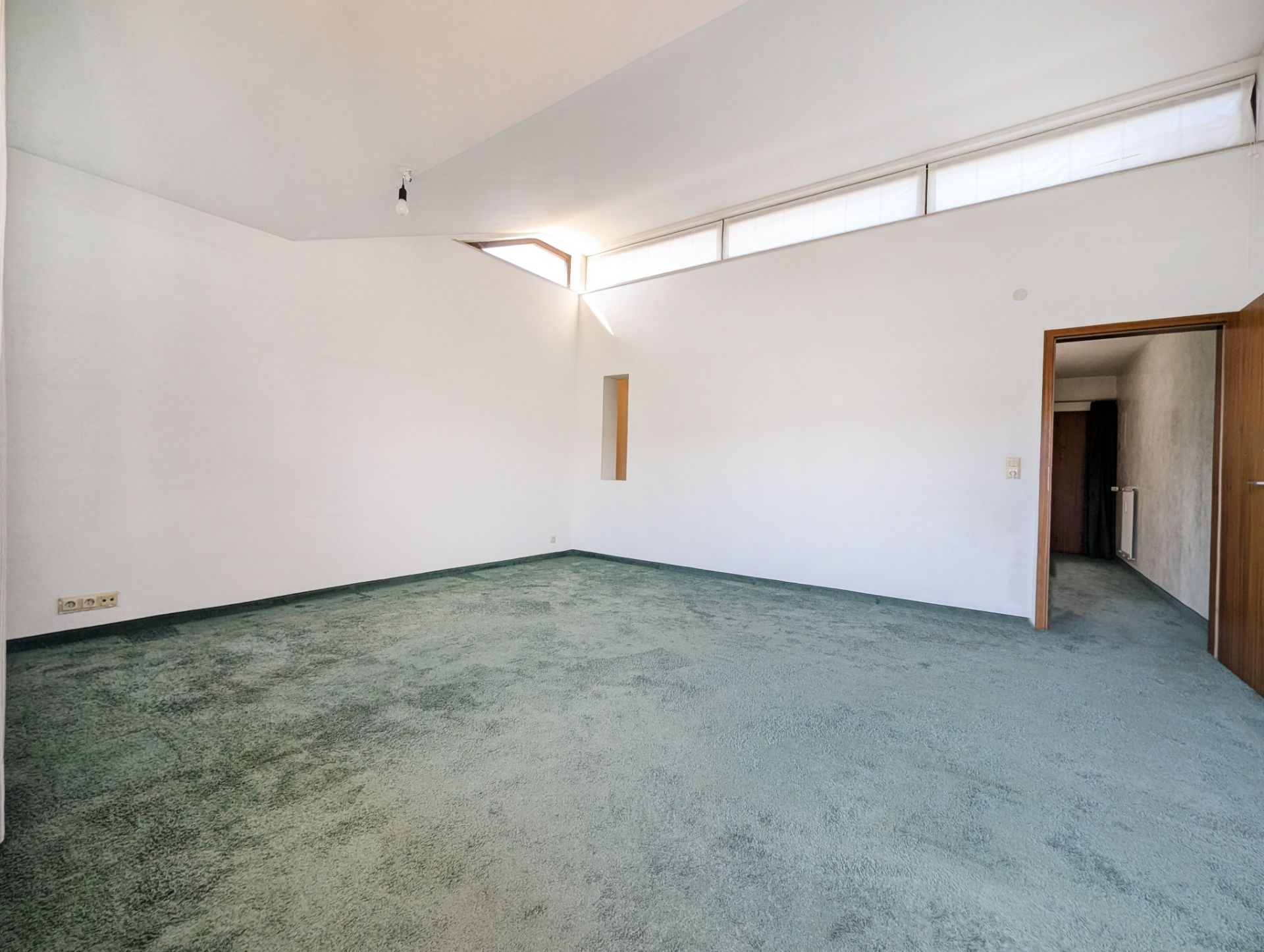 Leerer Raum mit grüner Auslegeware, weißen Wänden und langen Fenstern unter der Decke.