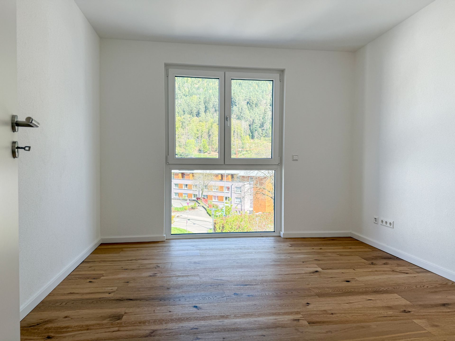 Leeres Zimmer mit Holzboden und großem Fenster, Blick nach draußen auf Bäume und Gebäude.