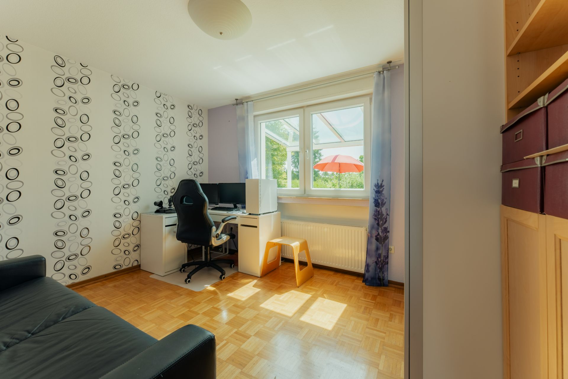 Helles Zimmer mit Schreibtisch, Stuhl, Sofa und großem Fenster mit Blick ins Grüne.