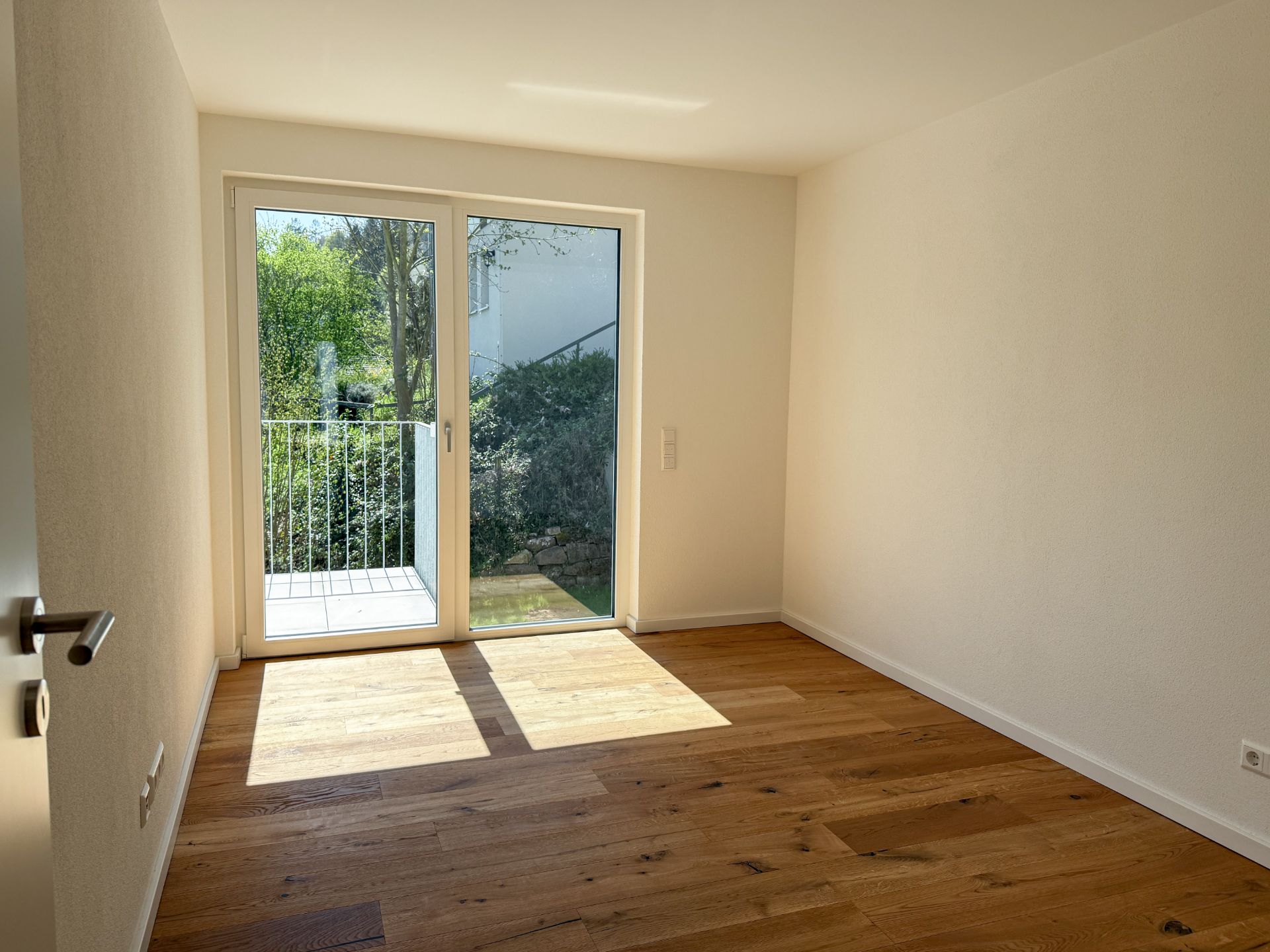 Helles Zimmer mit Holzboden und großer Glastür zur Terrasse, Blick ins Grüne.