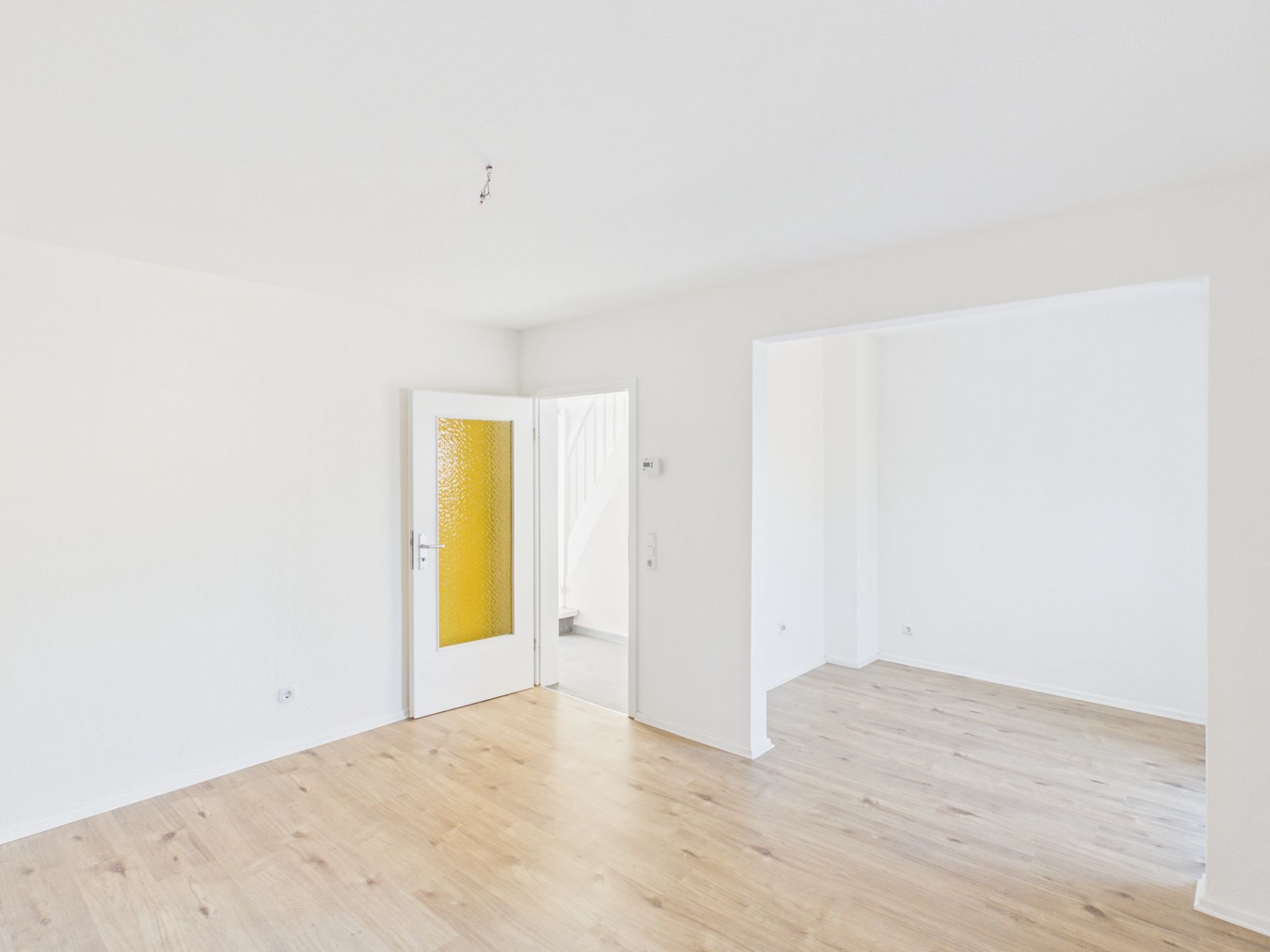 Heller, leerer Raum mit Holzboden, weißer Wand und Glastür mit gelbem Einsatz.