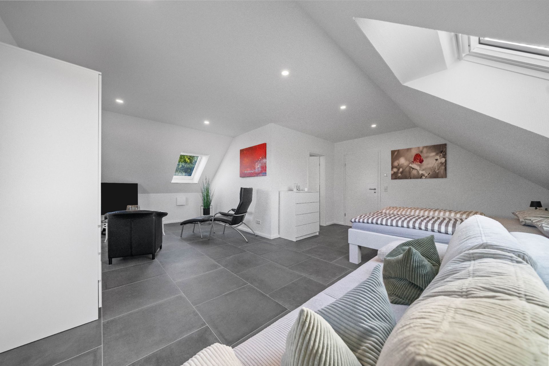 Helles Dachzimmer mit Sofa, Bett, Sesseln, Fernseher und Dachfenstern.