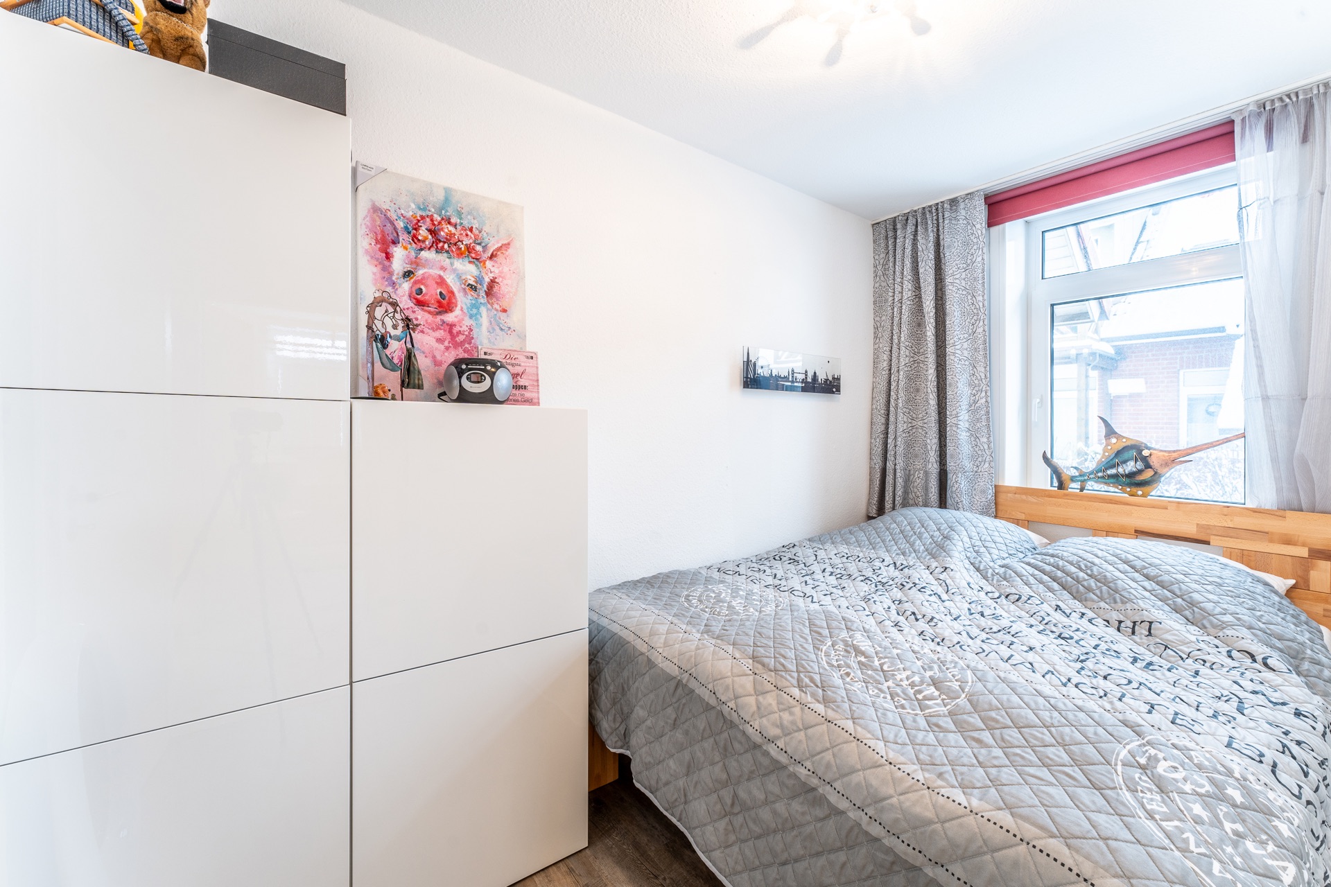 Helles Schlafzimmer mit grauem Bett, weißem Schrank und großem Fenster mit Vorhängen.