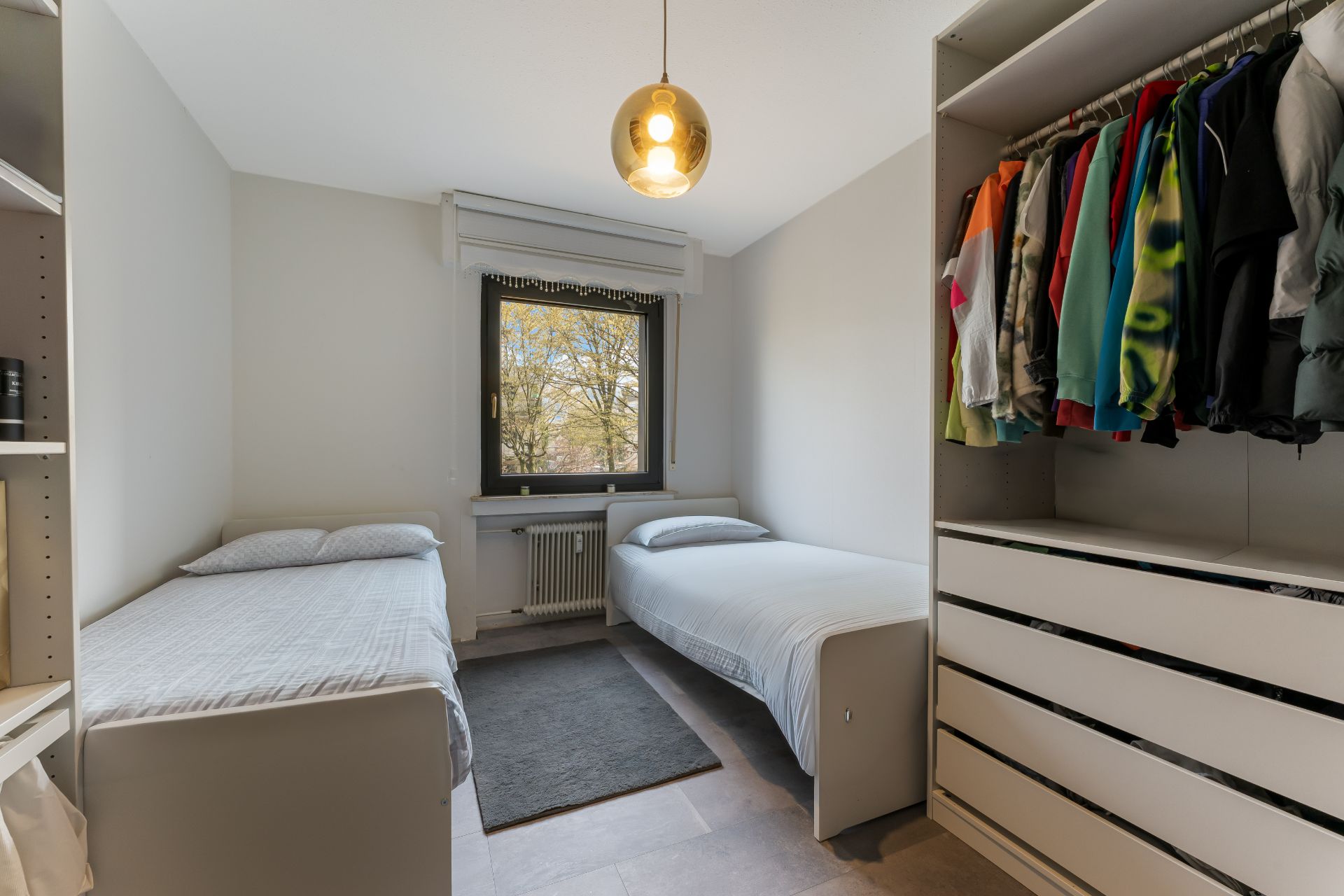 Zimmer mit zwei Einzelbetten, Fenster, Teppich und offenem Kleiderschrank mit Kleidung.