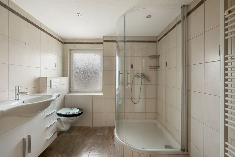 Helles Badezimmer mit großer Dusche, Waschbecken, Toilette und Fenster.