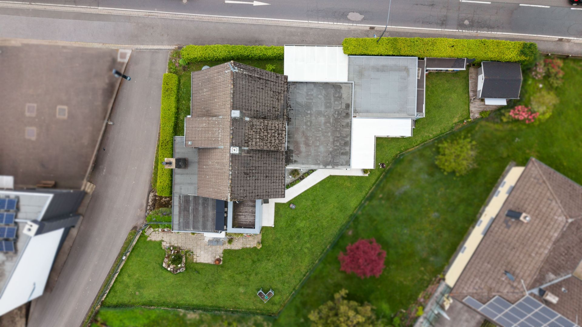 Luftaufnahme eines Hauses mit Garten und gepflasterter Auffahrt, umgeben von Straßen und Nachbargebäuden.